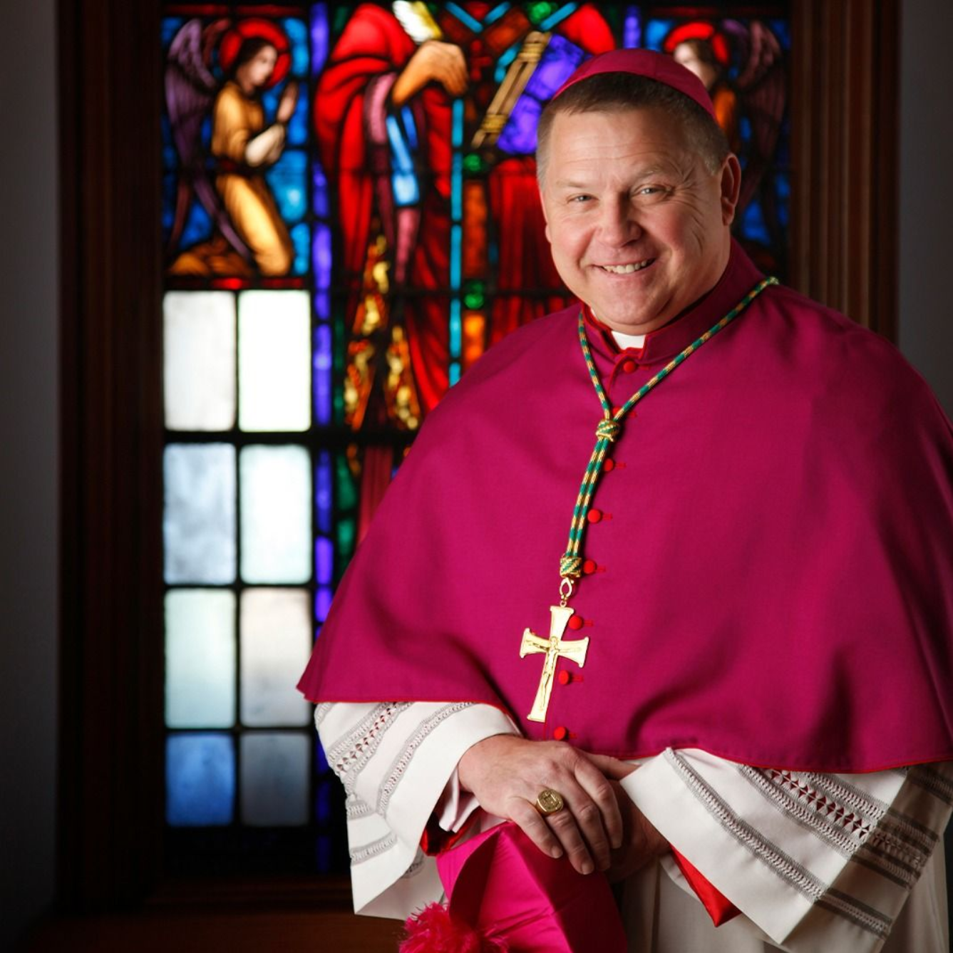 Archbishop Richard F. Stika in magenta robes smiles, holding a hat. Window with stained glass is in the background.