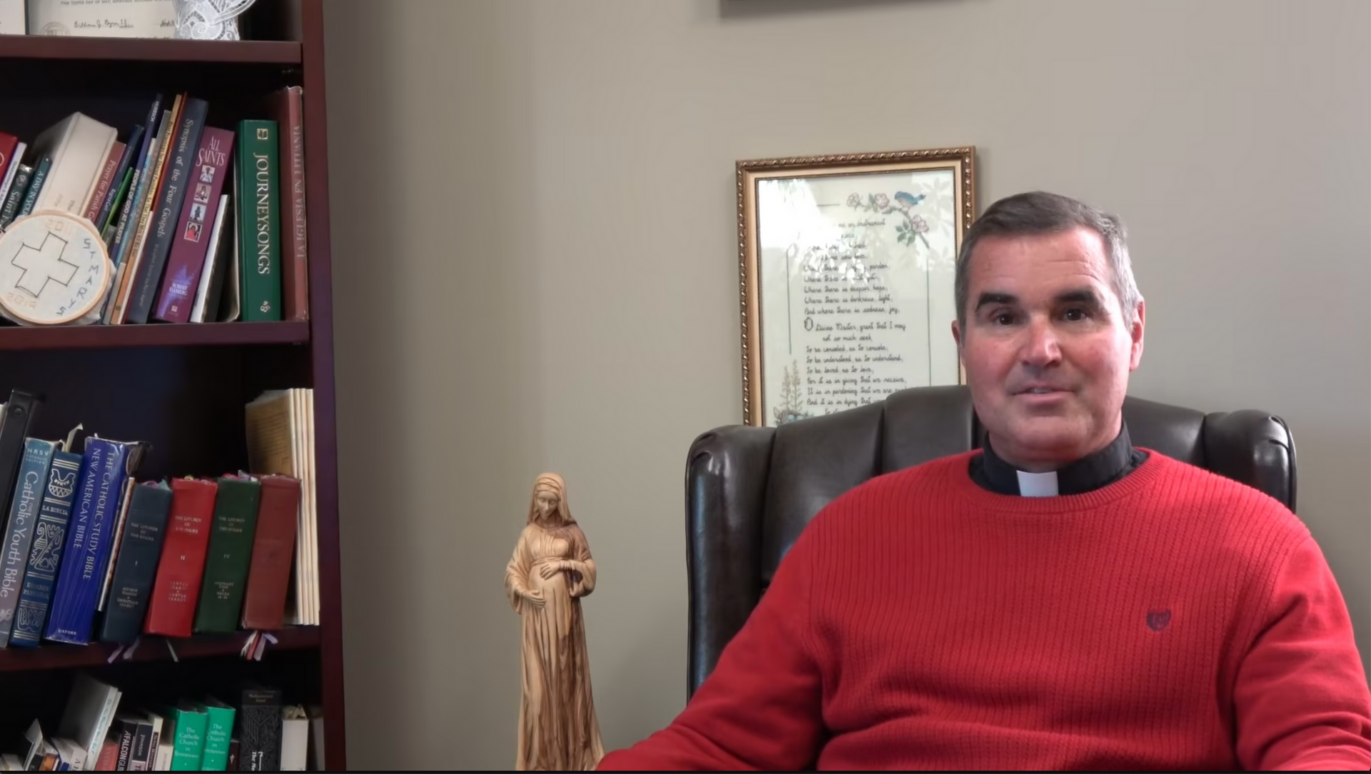 Clergyman in red sweater, seated at desk, looking at camera. Bookshelf and framed document visible.