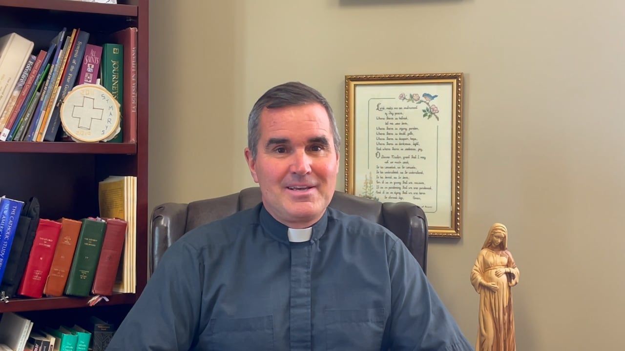 Man in clerical collar sits at a desk, speaking to the camera. Bookshelf and framed art are behind him.