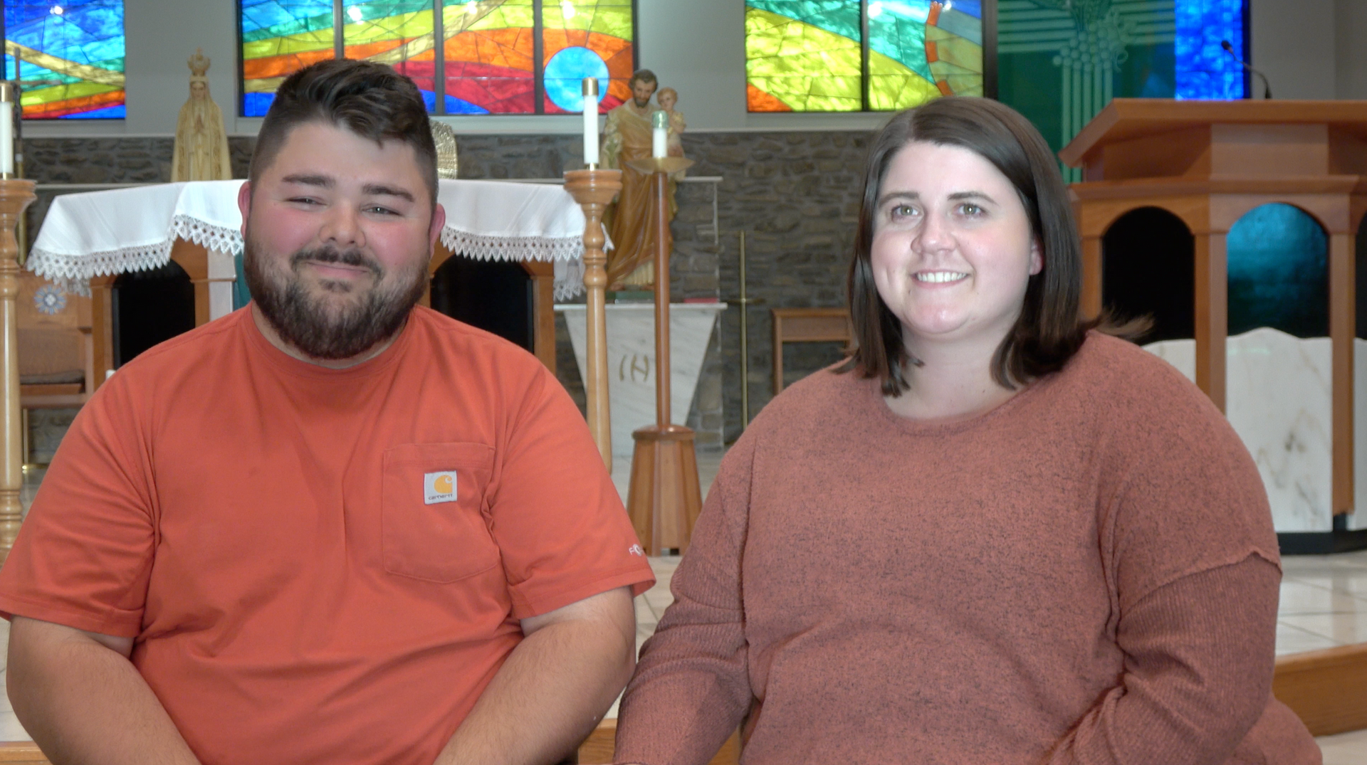 Couple in an orange shirt and sweater smiling in a church setting with stained-glass windows.