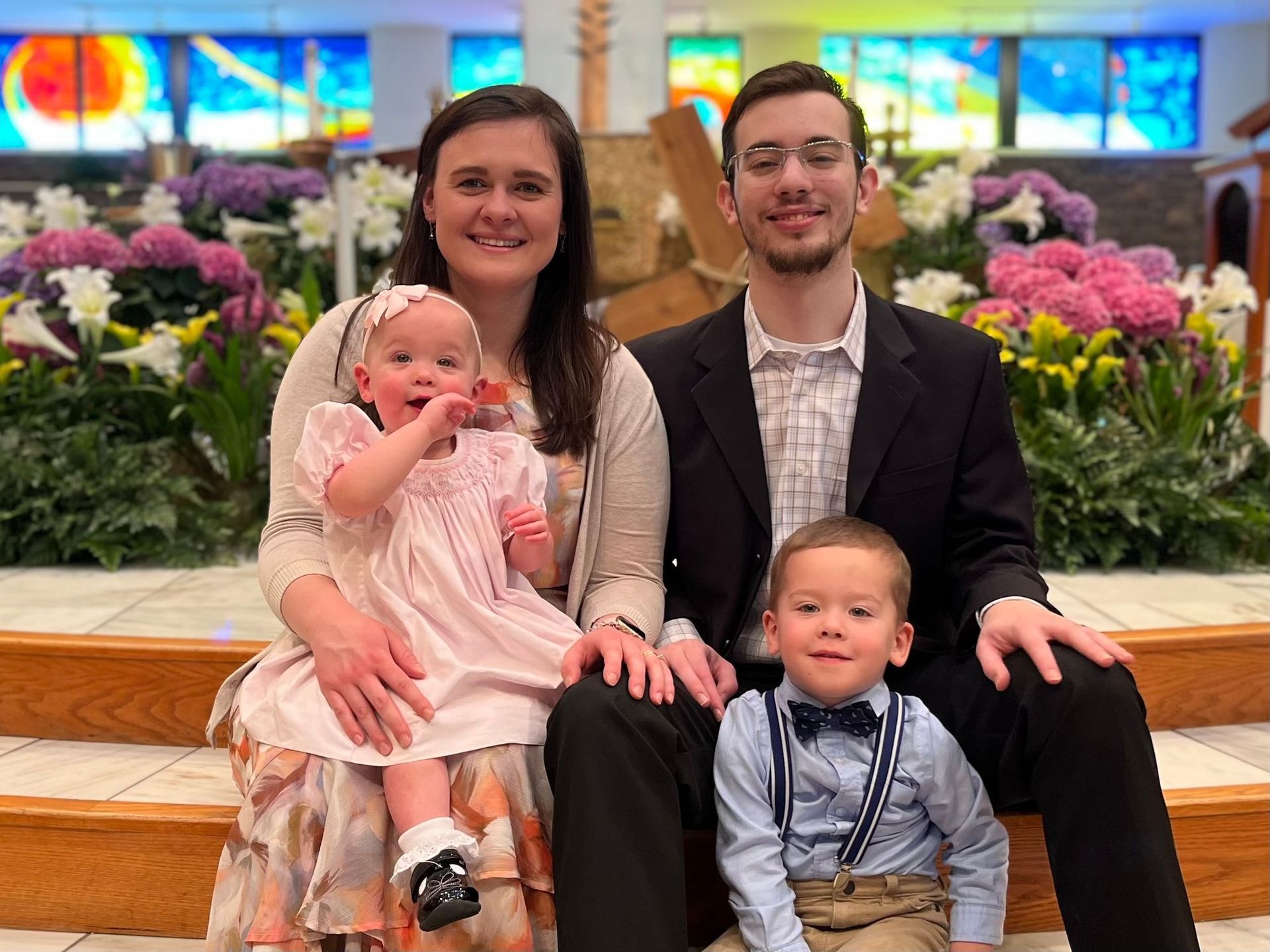 Family of four posing inside a church. Parents smiling with their two children, surrounded by flowers.