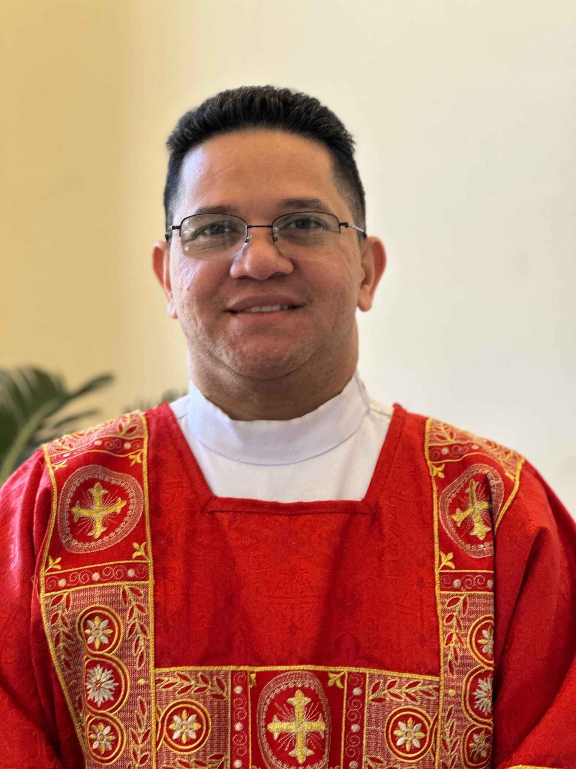 Deacon Renzo, soon to be Father Renzo smiling in red standing in front of a plant