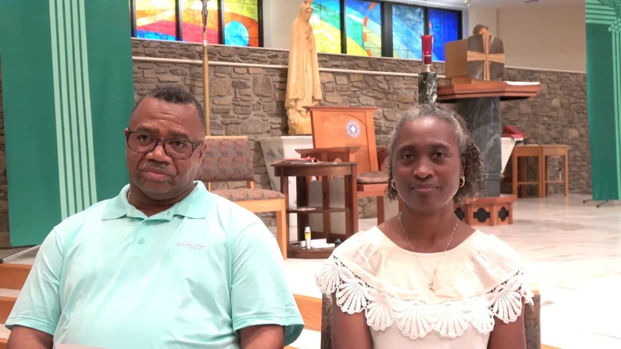 Man and woman seated, facing forward in church setting; man in blue shirt, woman in white top.
