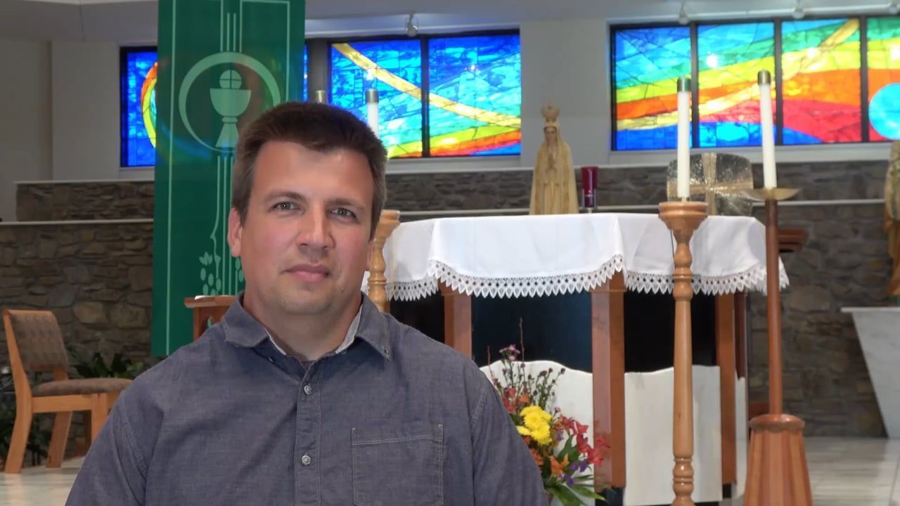 Man in front of a church altar, stained glass windows in background. He wears a button-down shirt.