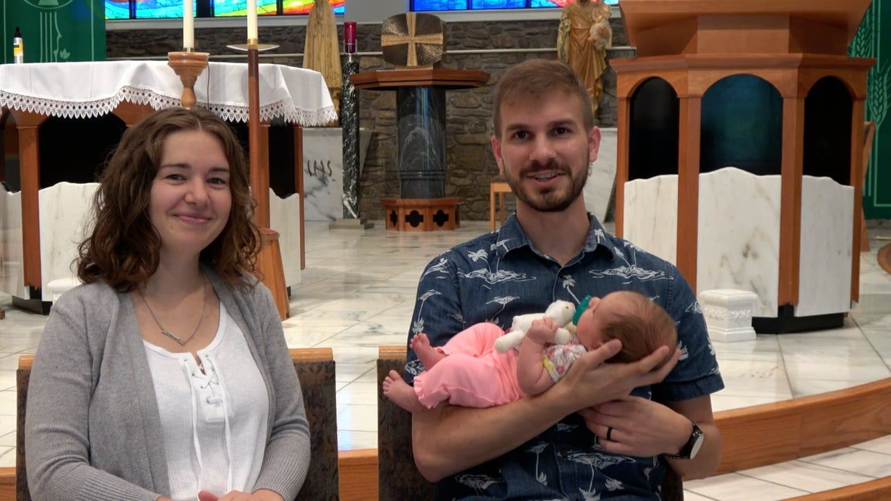 Parents holding baby in a church; woman in gray cardigan, man in blue shirt. Baby in pink.
