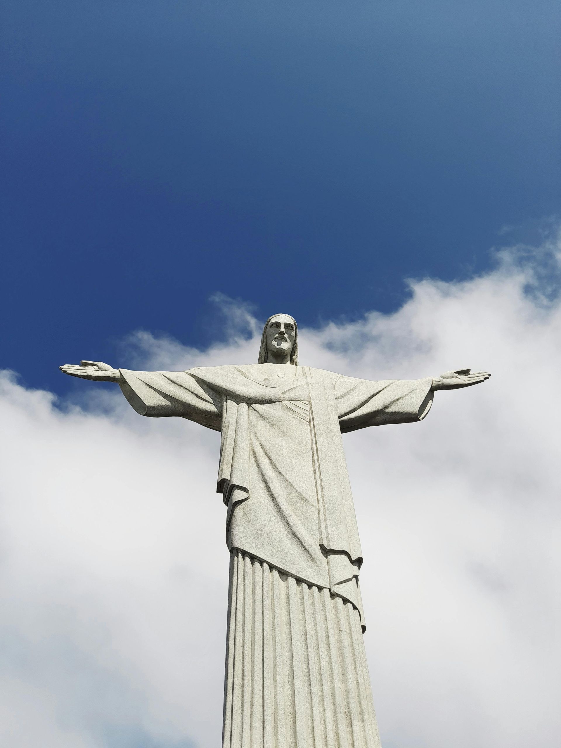 The Christ the Redeemer statue stands against a bright blue sky with white clouds, arms outstretched in a wide embrace.