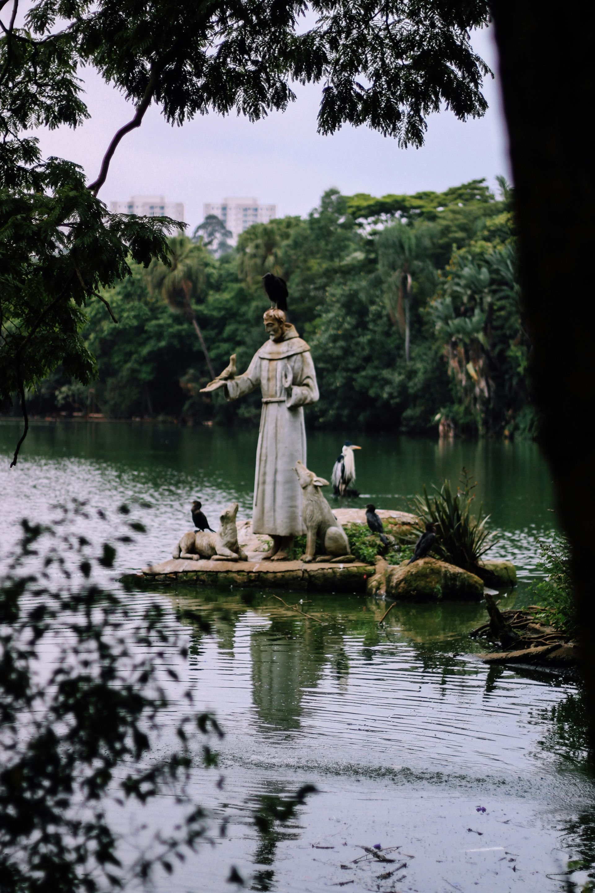 Statue of a man, surrounded by animals, stands in a lake. Green trees and buildings are in the background.