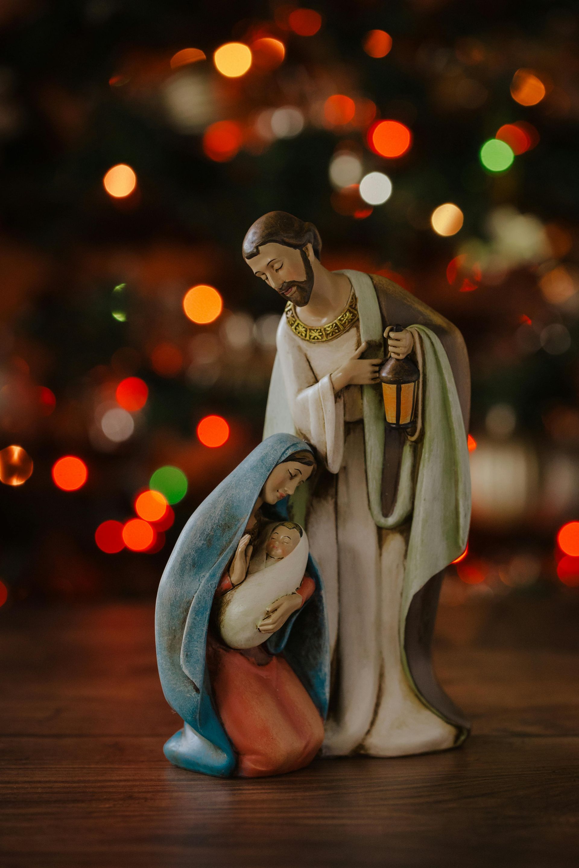 Nativity scene figurine: Mary and Joseph with baby Jesus, with Christmas tree lights blurred in background.