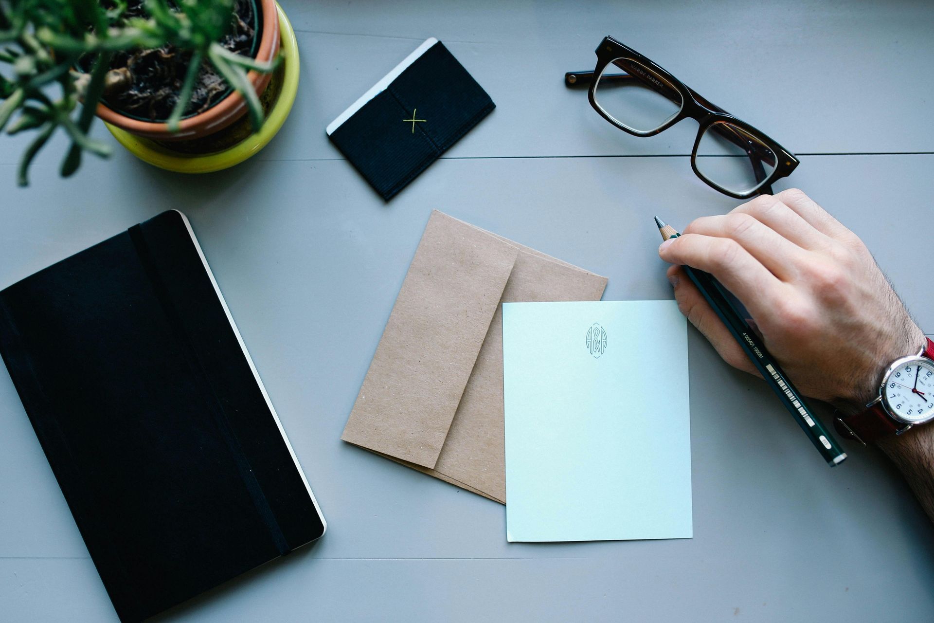 Person writing on a light blue paper on a gray desk, alongside a notebook, glasses, and an envelope.