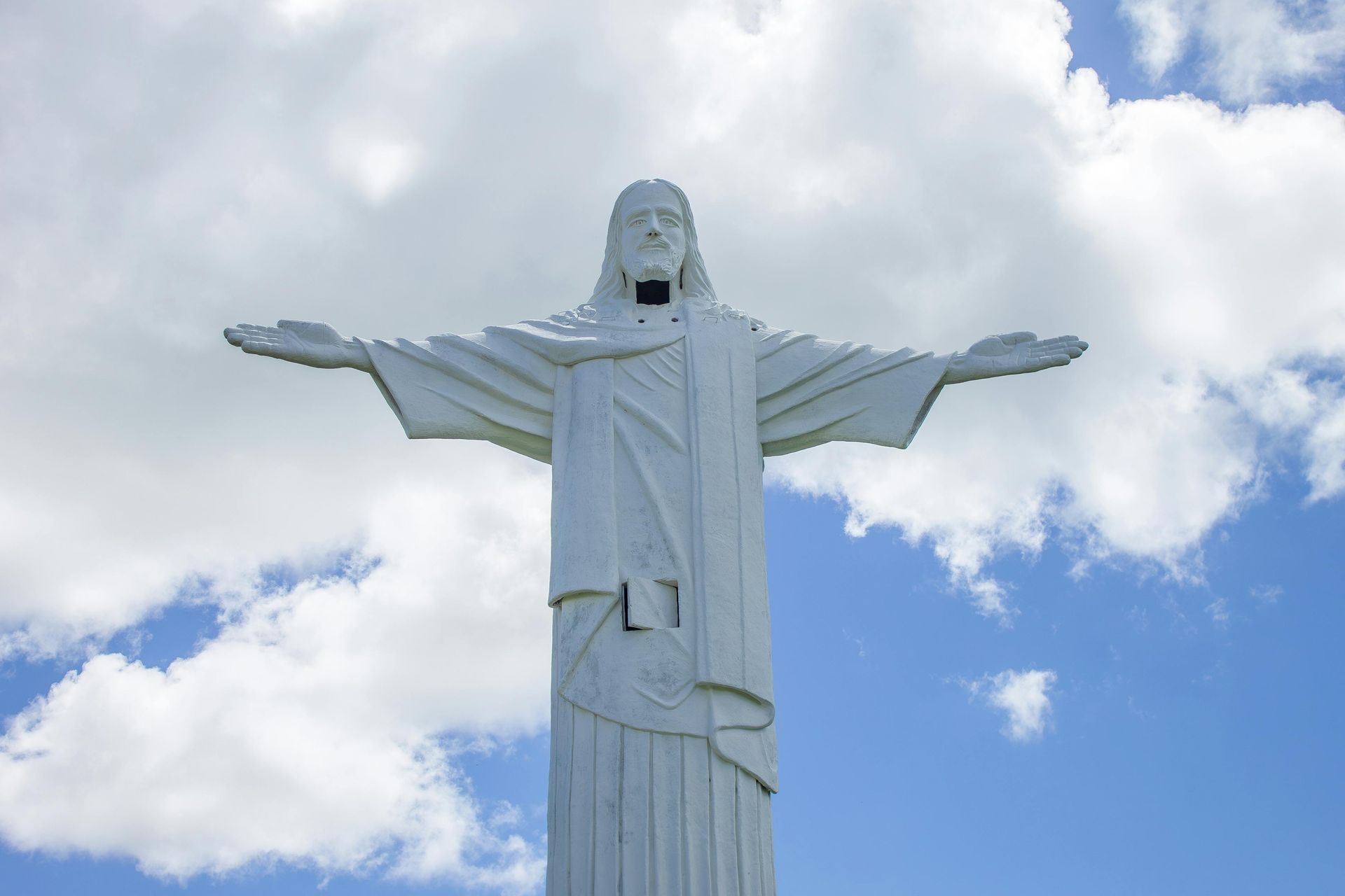 A white, towering statue of Jesus Christ with arms outstretched against a bright blue sky with scattered white clouds.