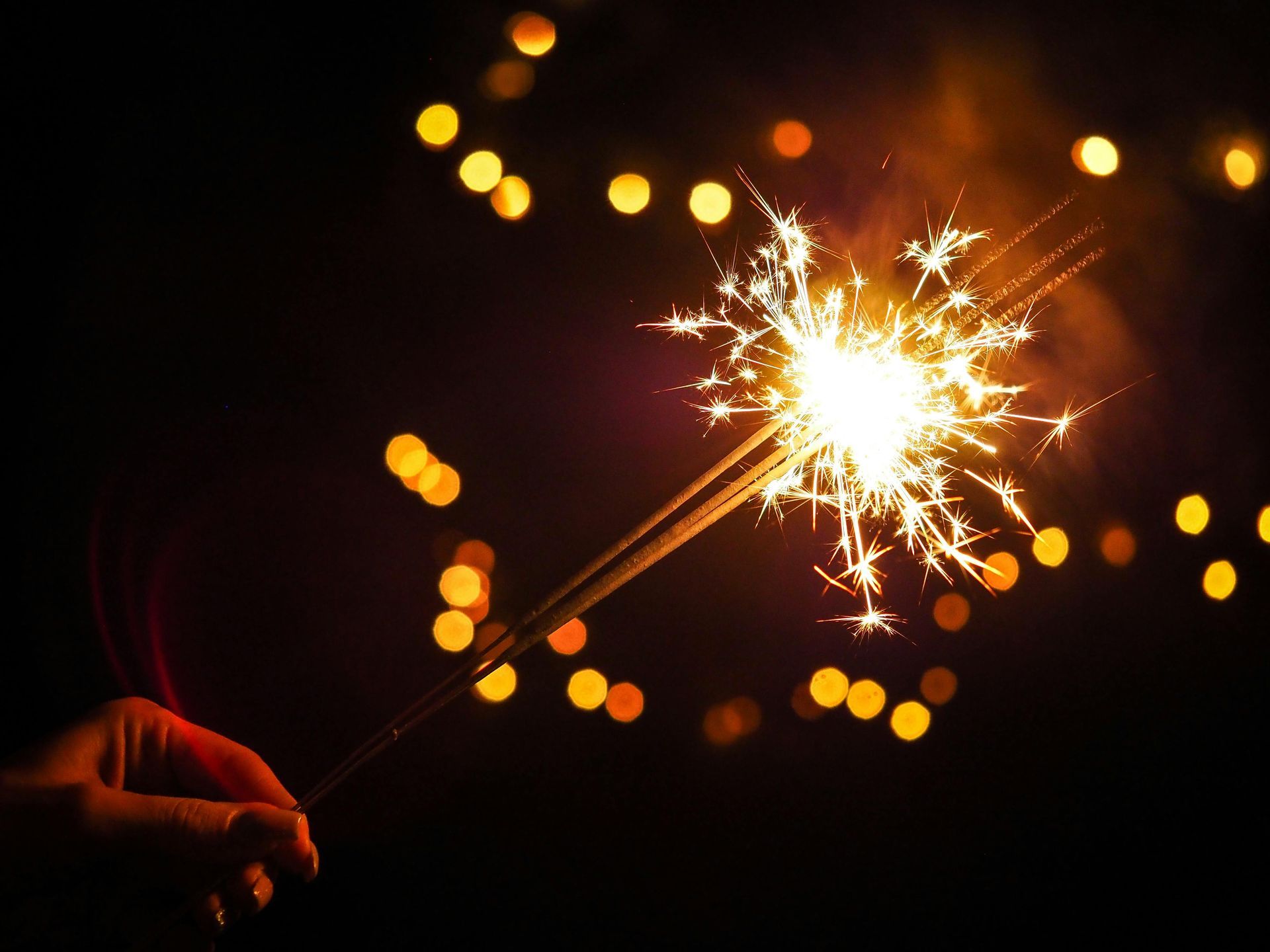 Hand holding a lit sparkler, with sparks radiating outwards. Bokeh lights in the dark background.