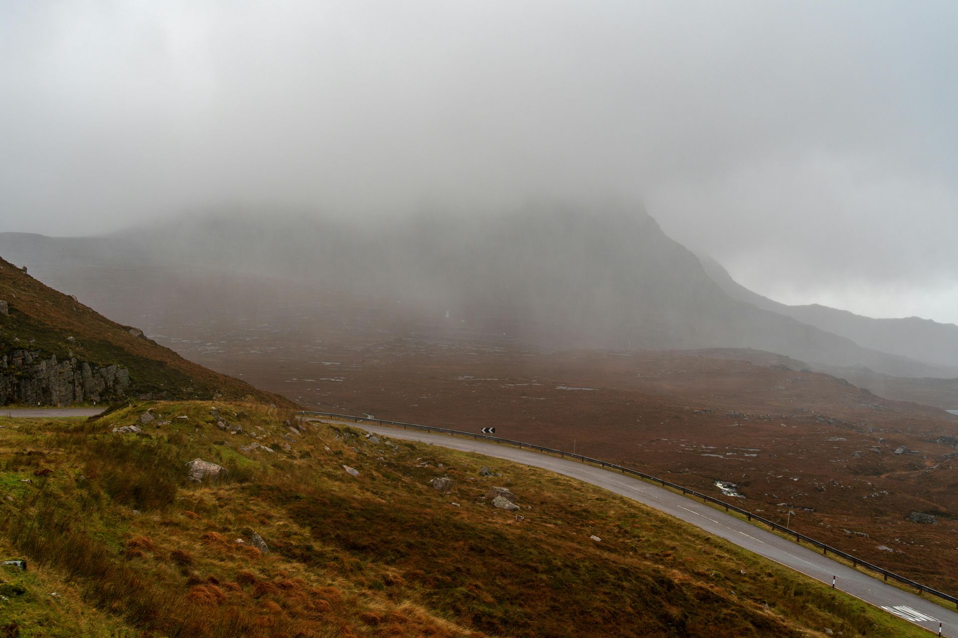 Winding road through a rugged, brown landscape shrouded in mist. A mountain peak is partially visible.