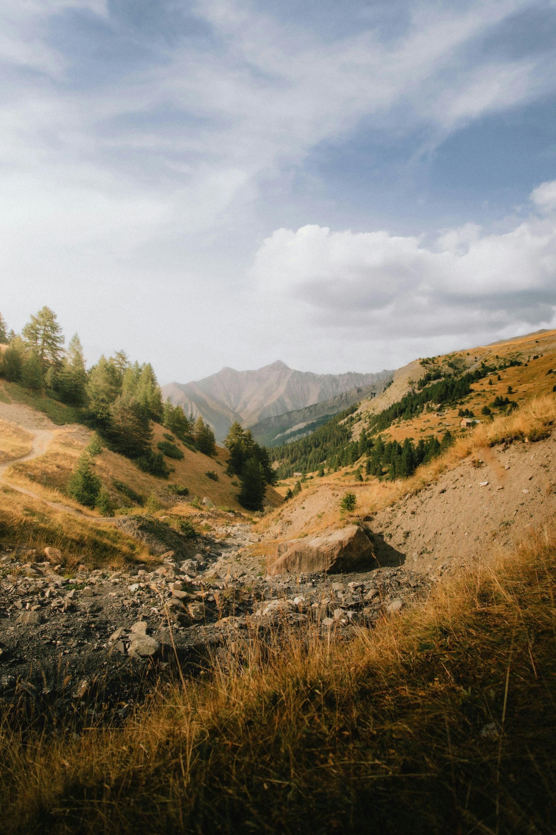 Mountain valley landscape with dry vegetation, sparse trees, and a cloudy blue sky.