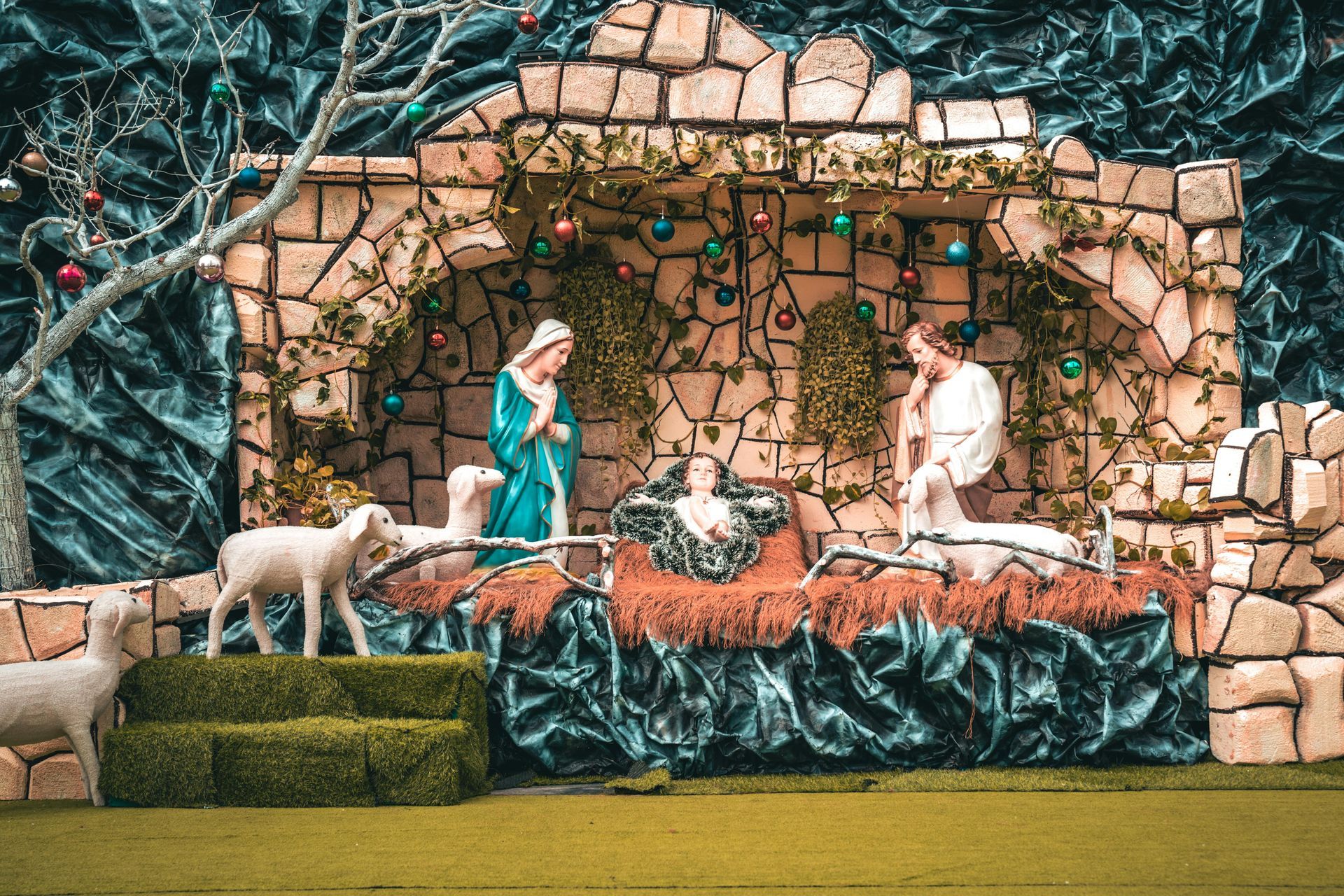 Nativity scene: Mary and Joseph beside baby Jesus in a manger, with sheep and decorative stone building.