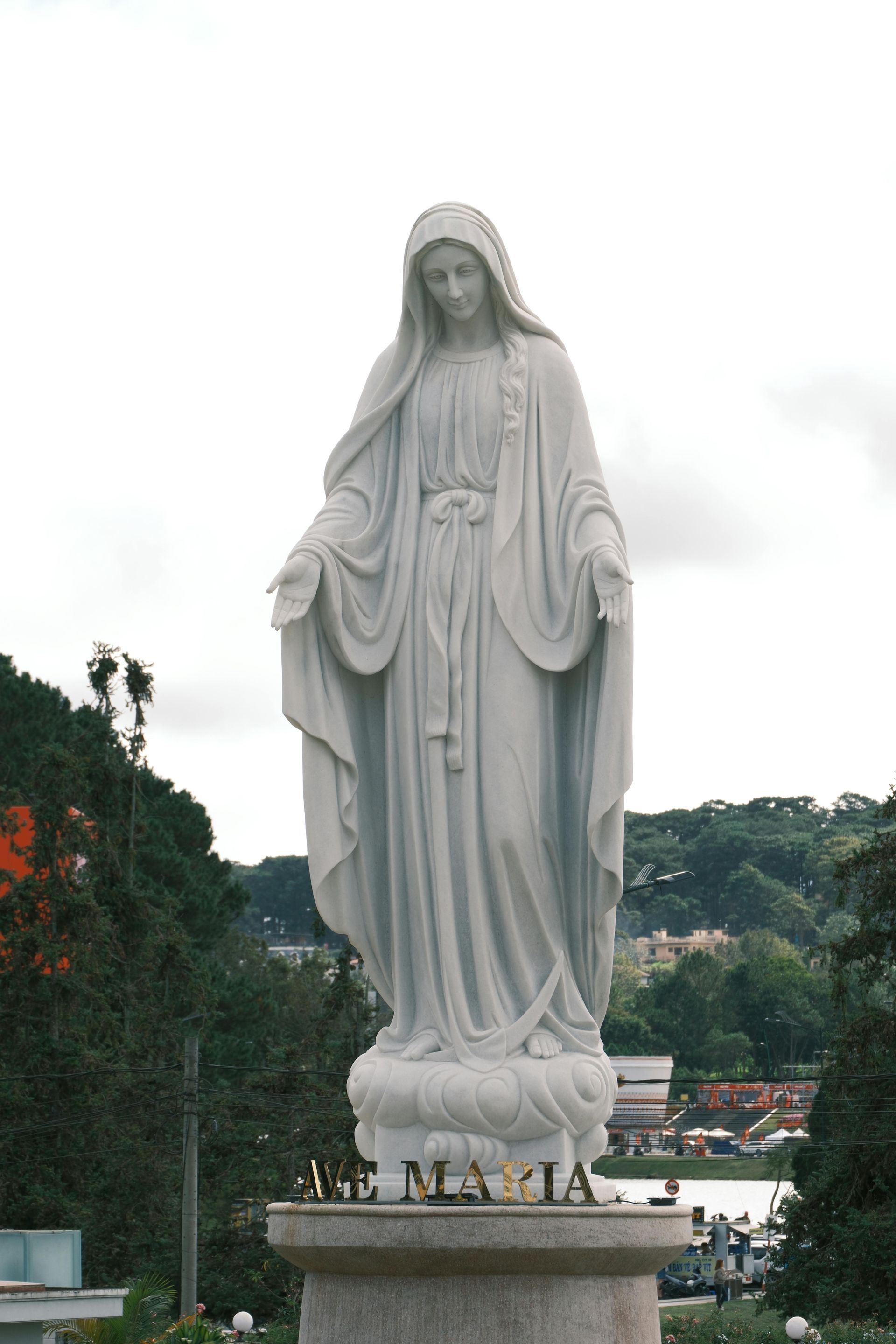 Statue of Virgin Mary, white stone, standing, arms open, on a pedestal. 