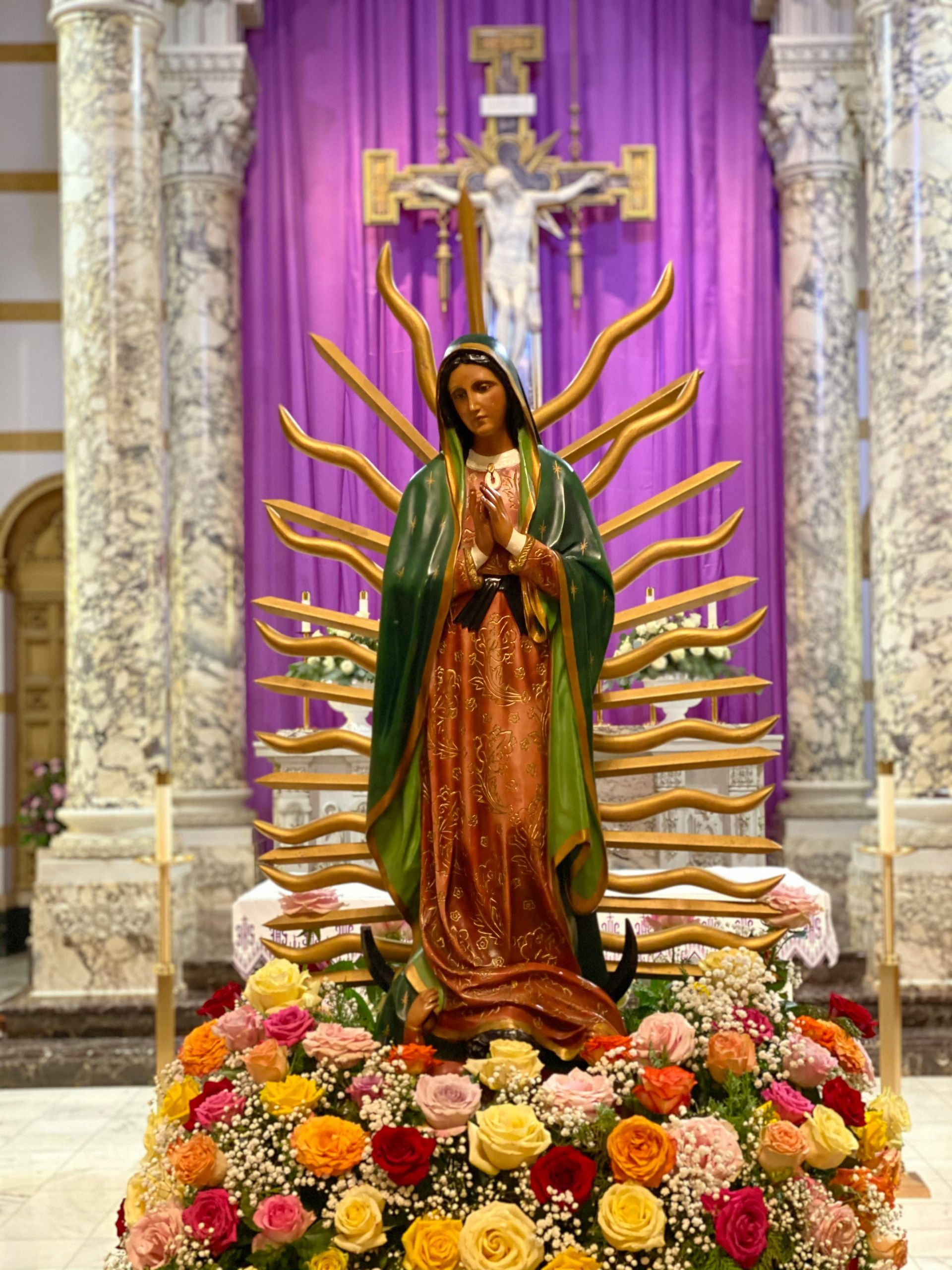 Statue of the Virgin Mary, surrounded by floral display, in front of a purple draped altar with a crucifix.