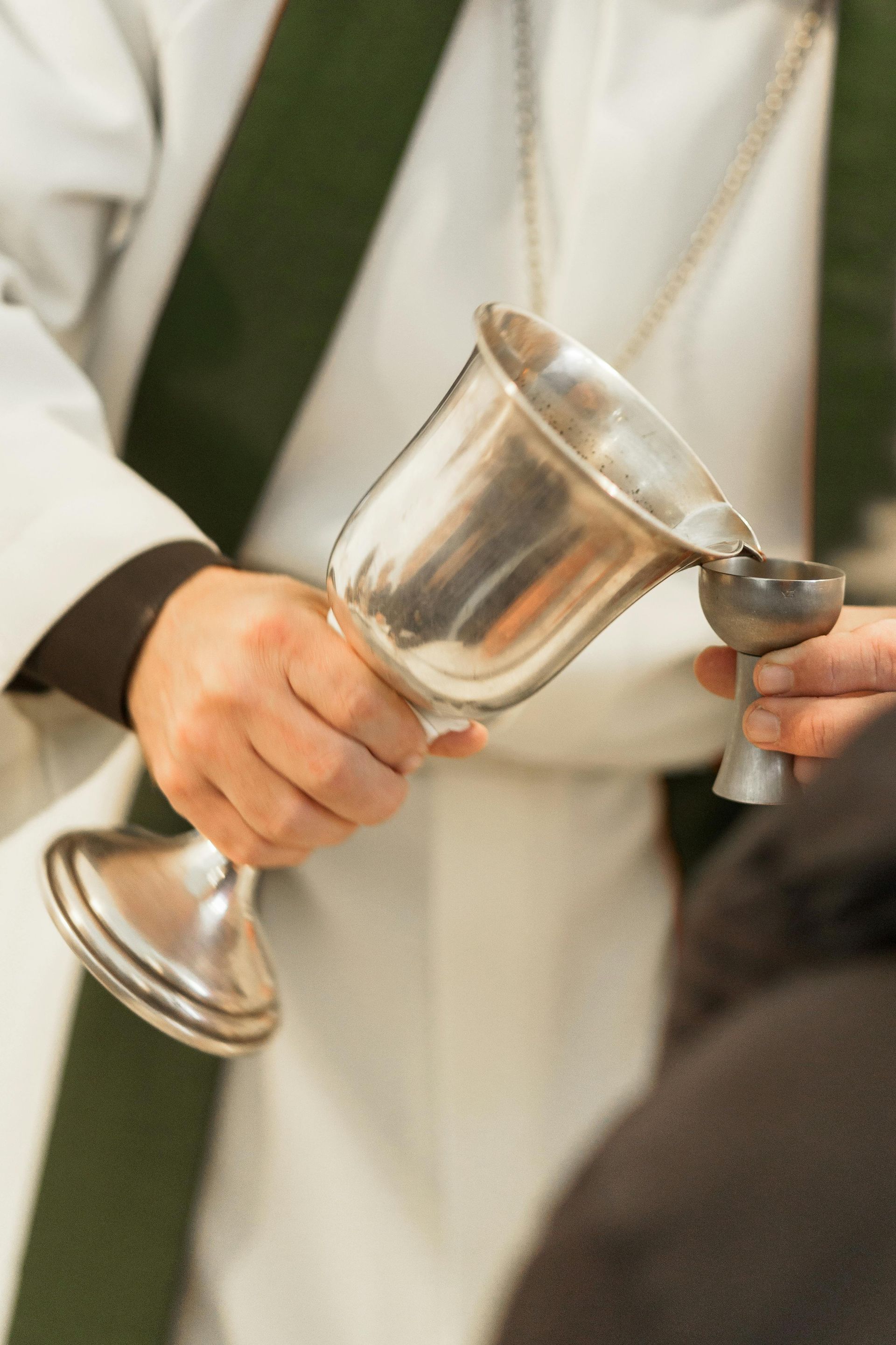 A priest in white and green vestments pours wine from a chalice into a small cup during a religious service.