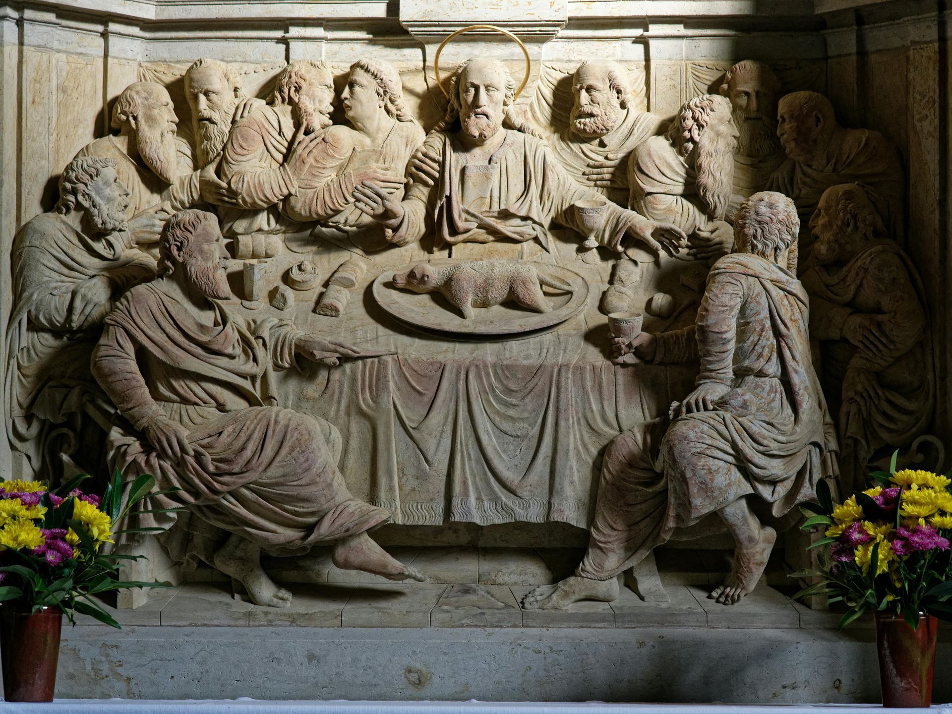 Stone relief sculpture of The Last Supper featuring Jesus and apostles seated at a table with food, with floral vases.