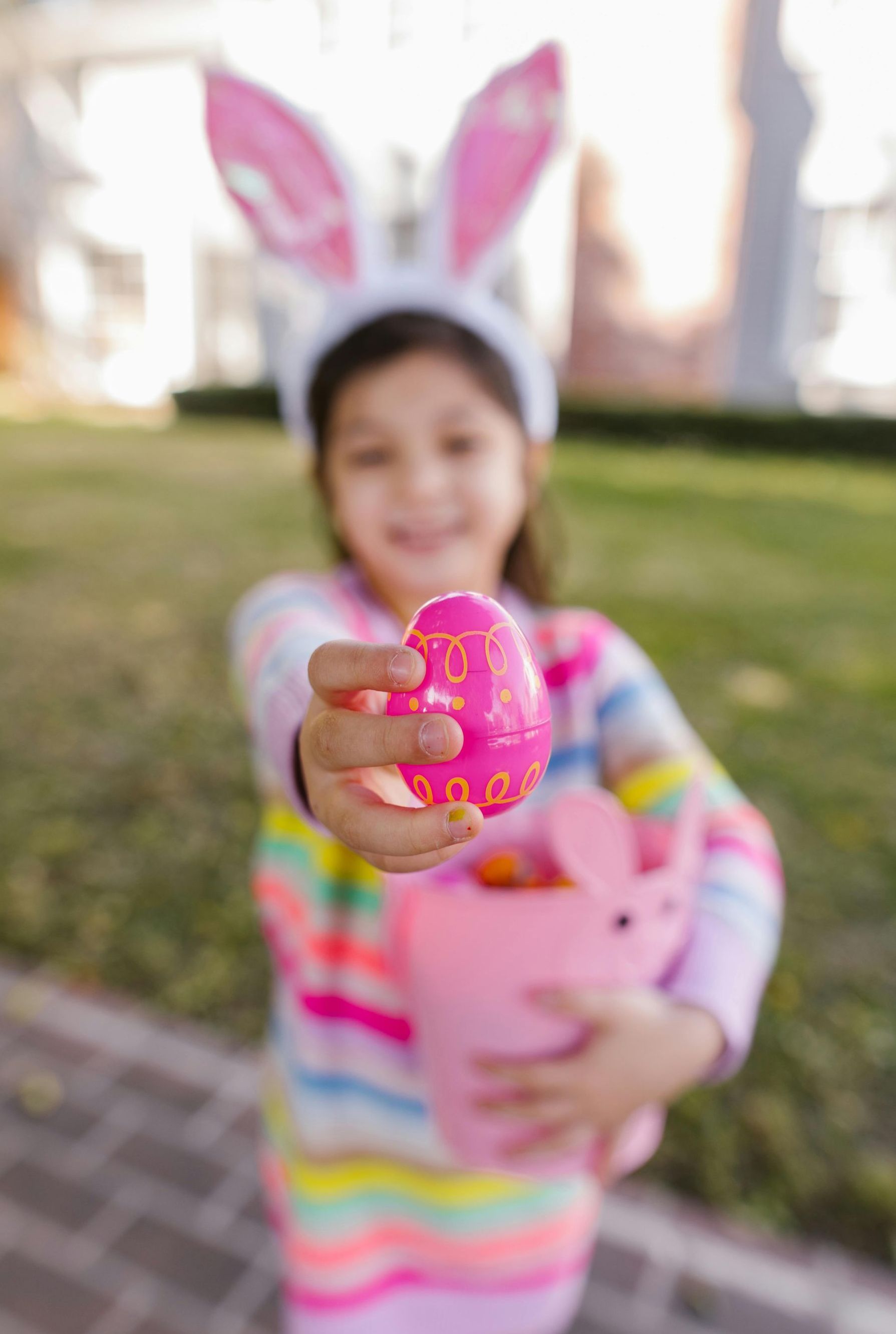 A little girl wearing bunny ears is holding a pink easter egg.