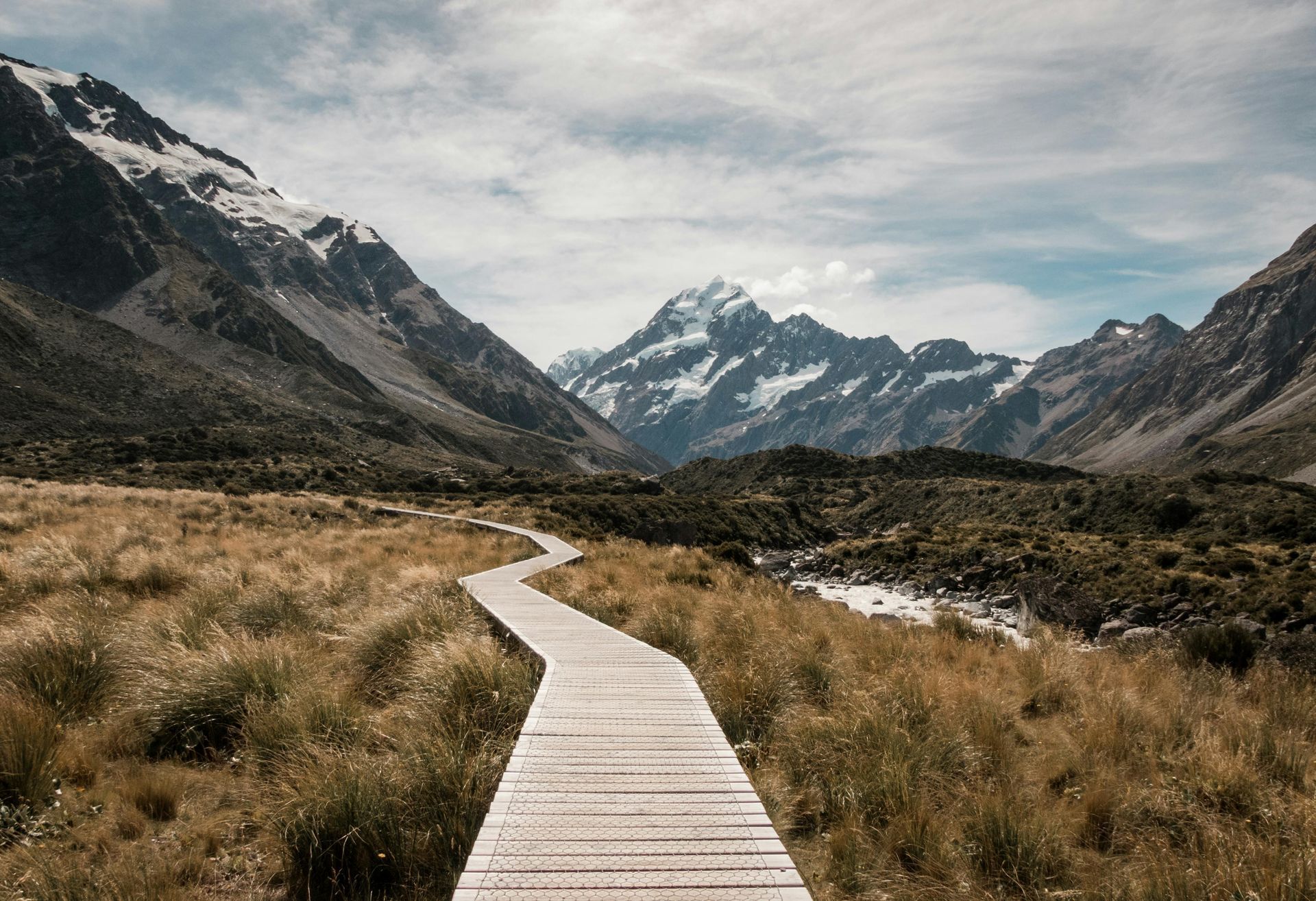 Wooden walkway through a grassy field, leading toward snow-capped mountains under a cloudy sky.