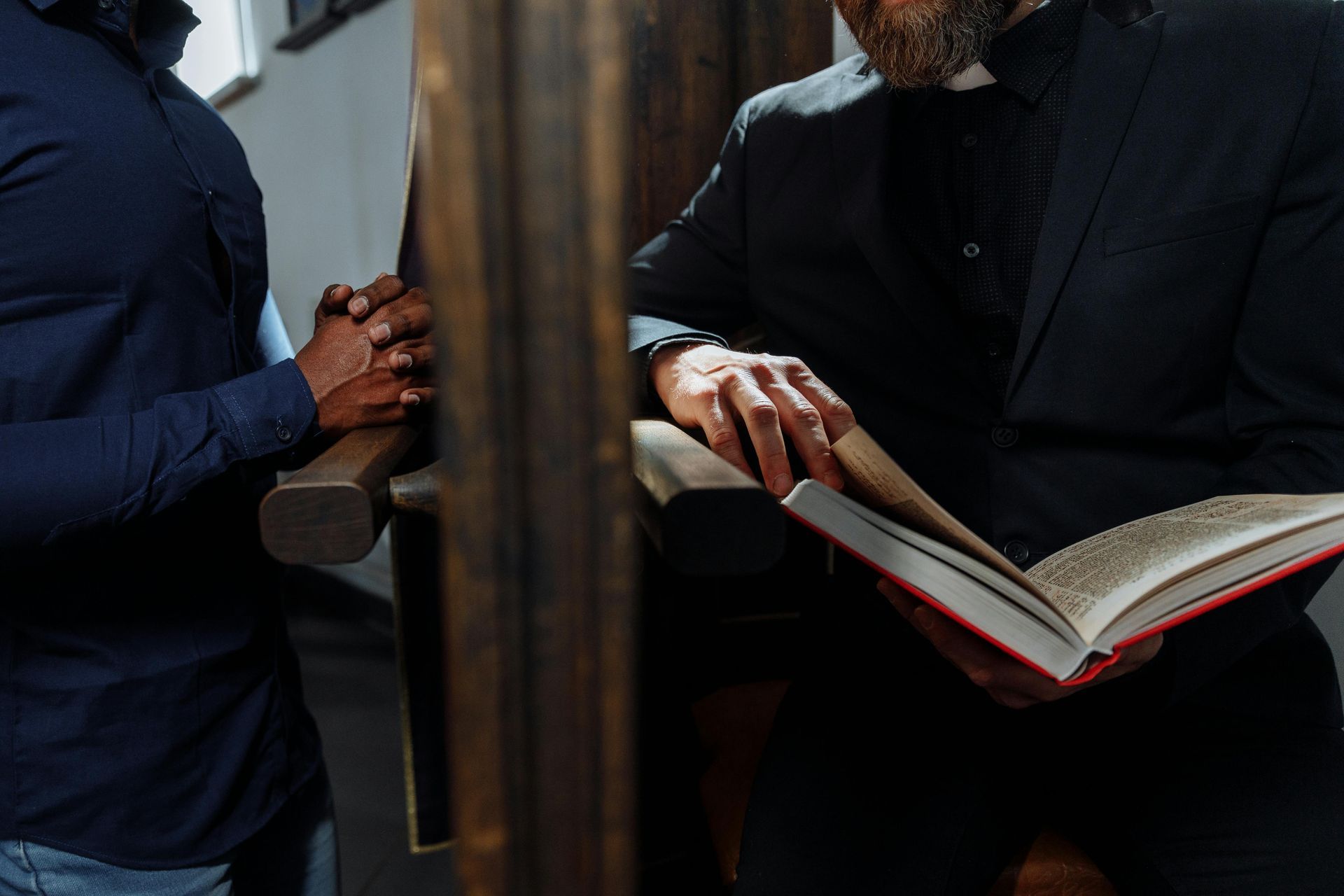 A priest in a dimly lit confessional booth, reading a book with sunlight casting shadows.