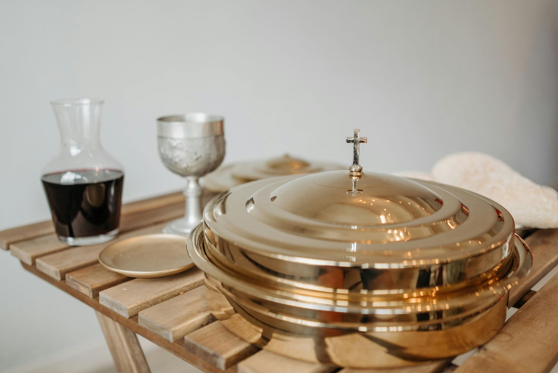 A gold-plated communion set with a chalice, carafe of wine, and covered bread plate resting on a wooden slatted table.