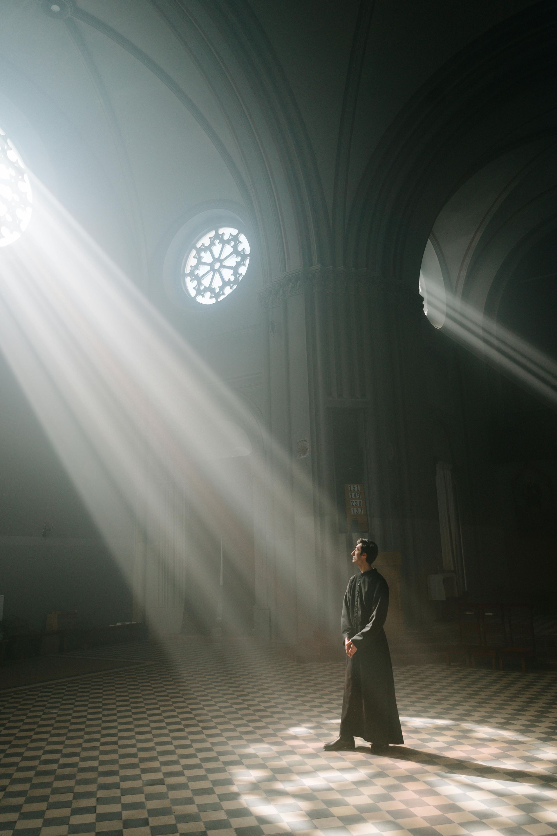 A person in dark robes stands in a cathedral, illuminated by a ray of light.