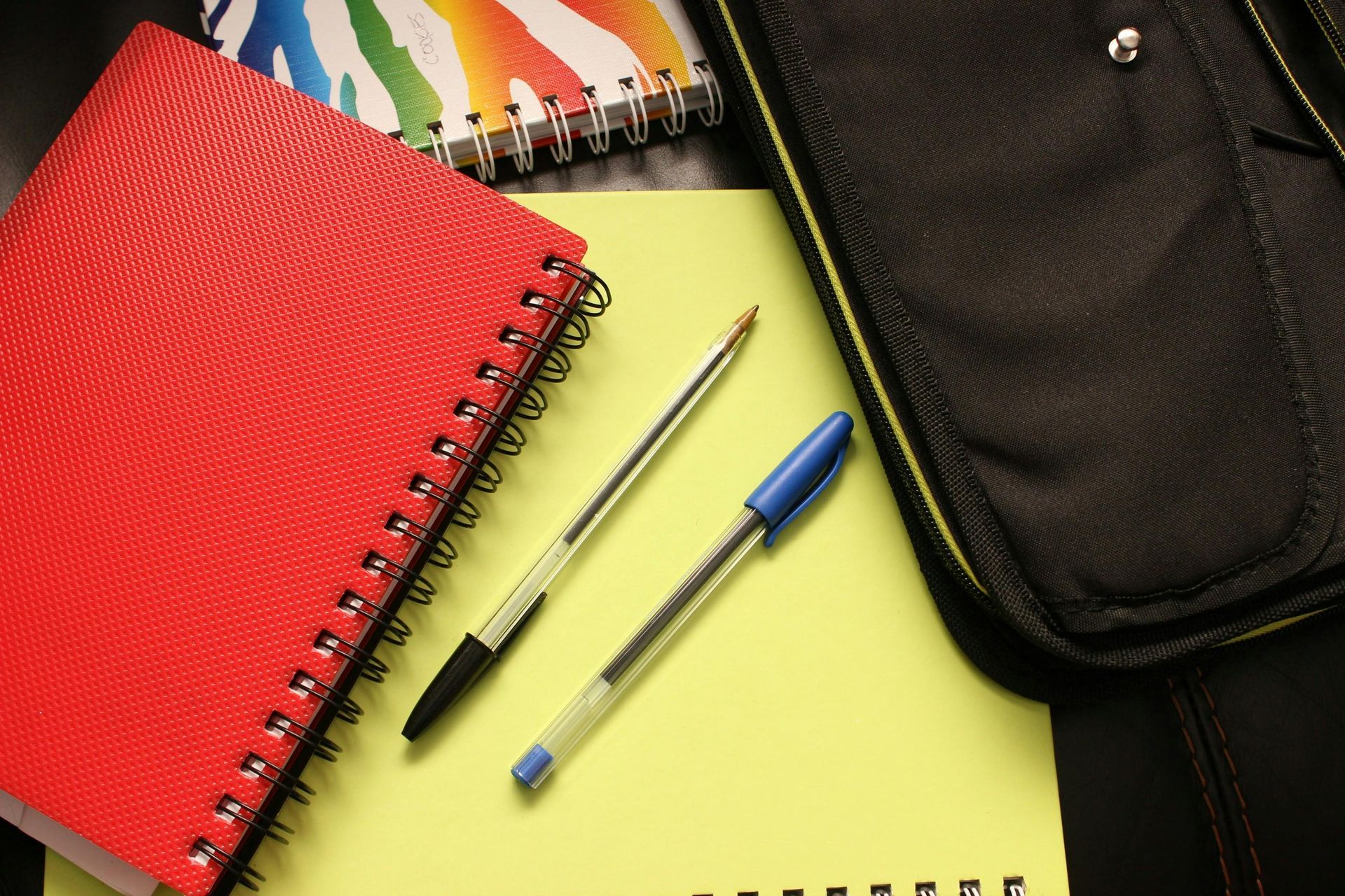 Red spiral notebook, pens, and a black bag on a yellow pad of paper. Other notebooks are visible.