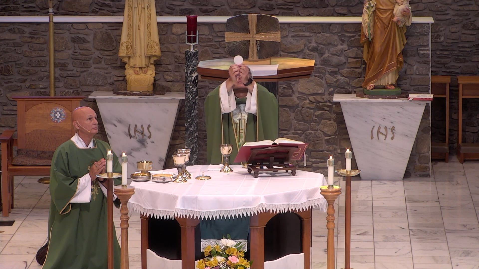 Fr consecrates the Eucharist at an altar, with an acolyte, candles, and church setting.
