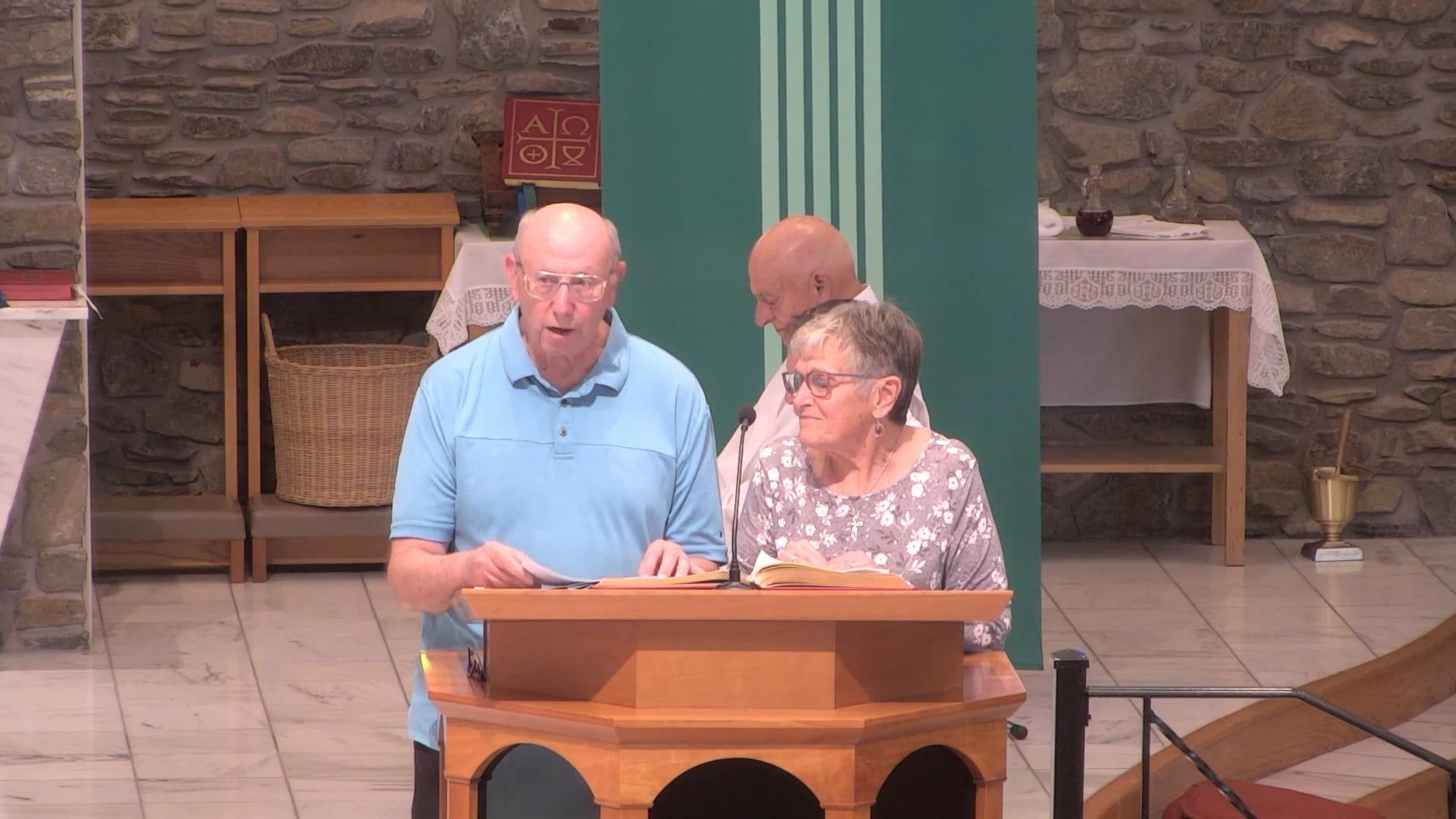 Two people at a wooden lectern in a church, speaking. A third person stands behind them.