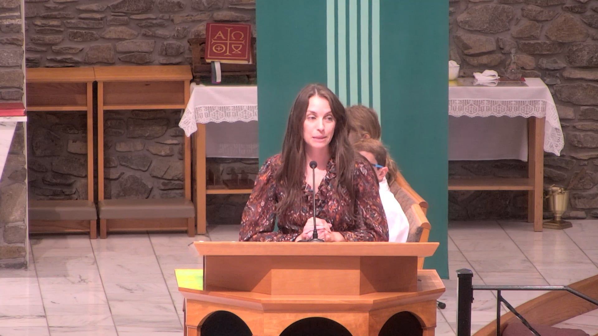 Woman speaking at a wooden podium in a church, with a green backdrop and stone walls.