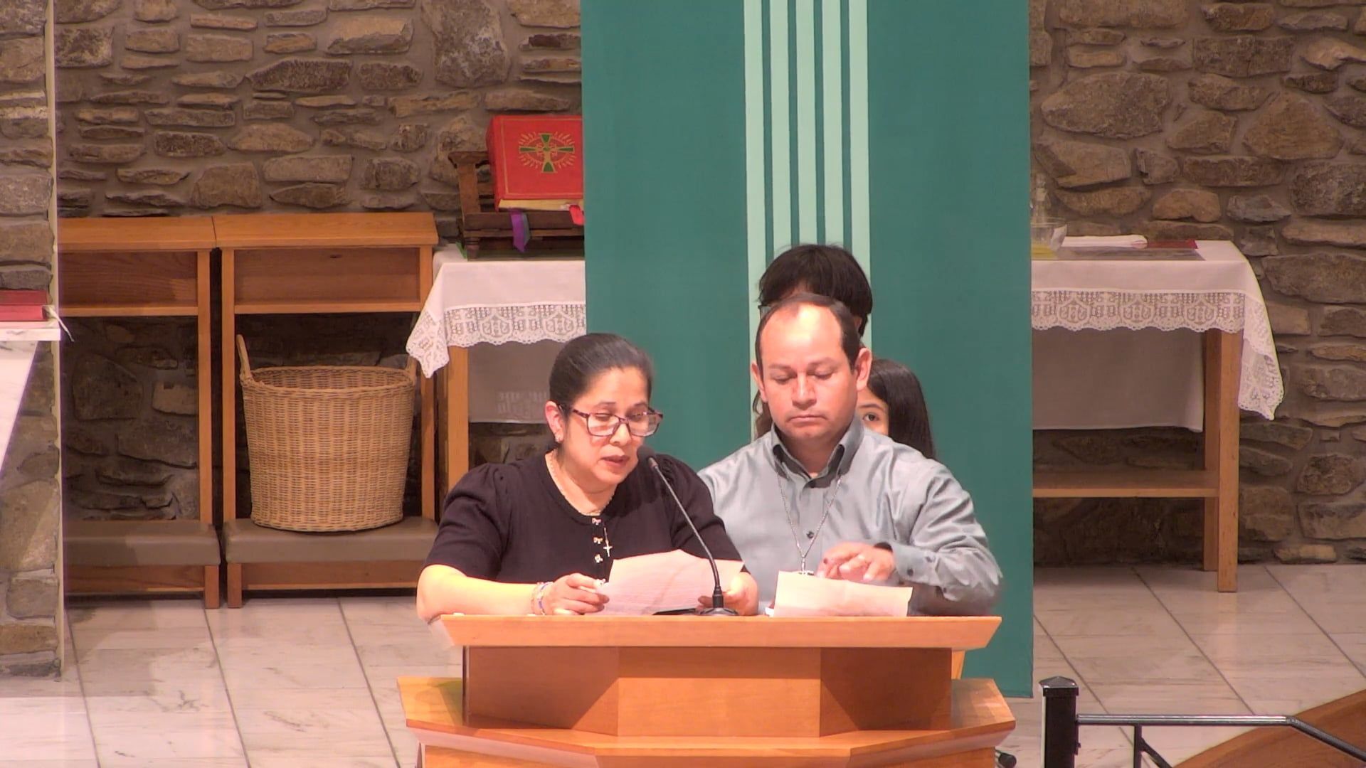 Two people reading at a podium in a church, green backdrop, stone walls.