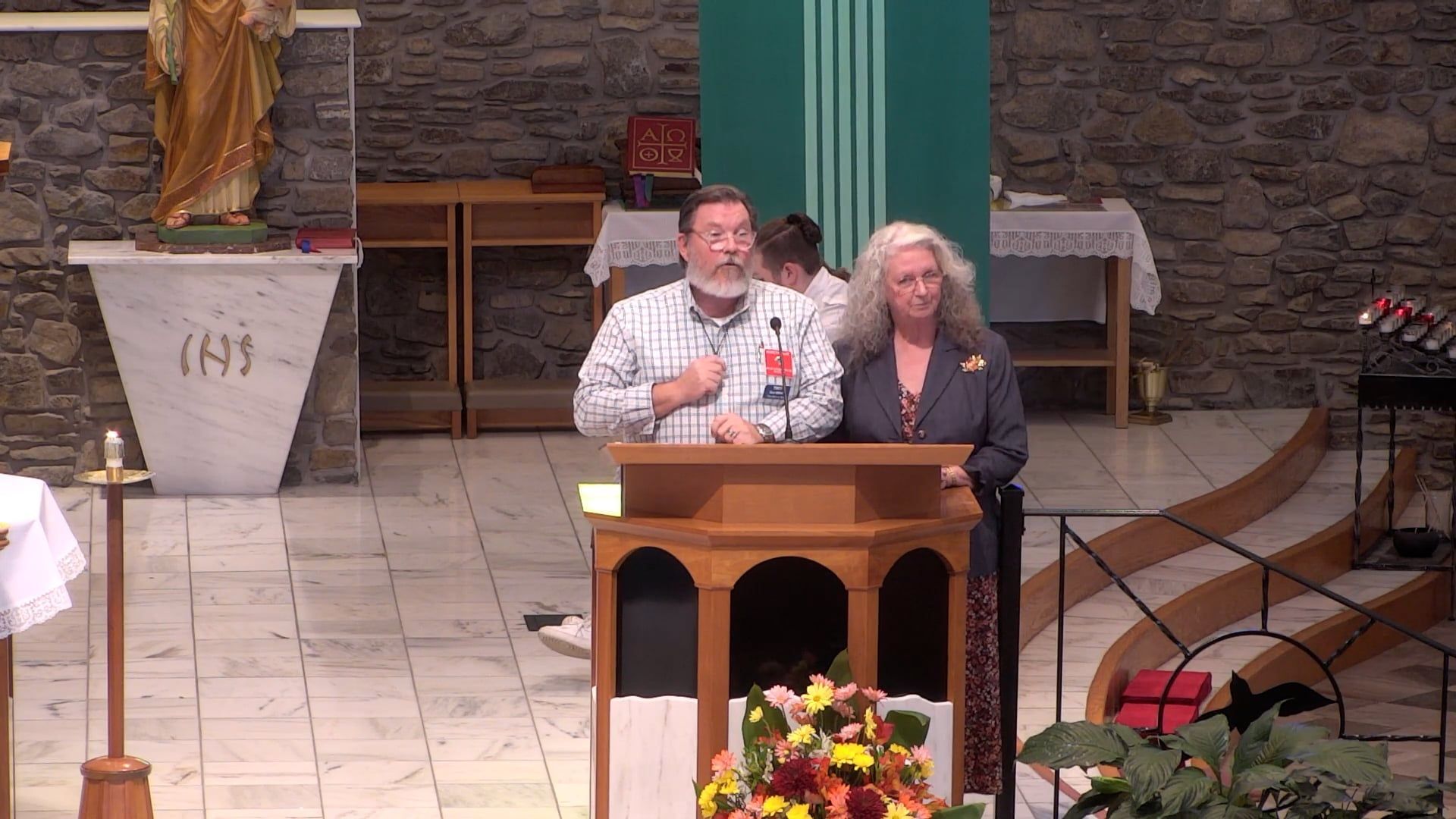 Two people speaking at a wooden podium in a church. A statue of Jesus is visible.