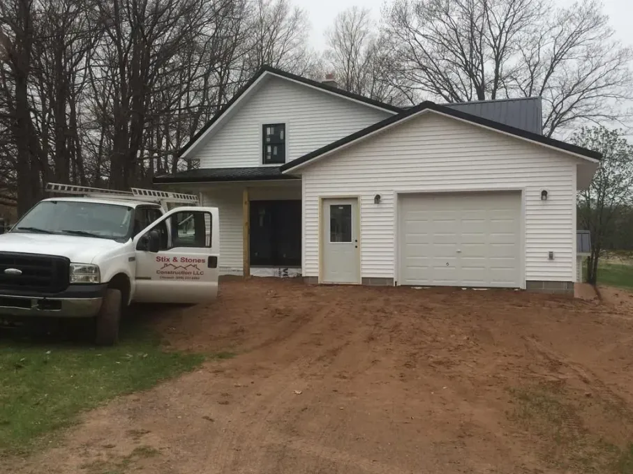 White house with a garage and parked work truck on a dirt driveway, surrounded by bare trees.