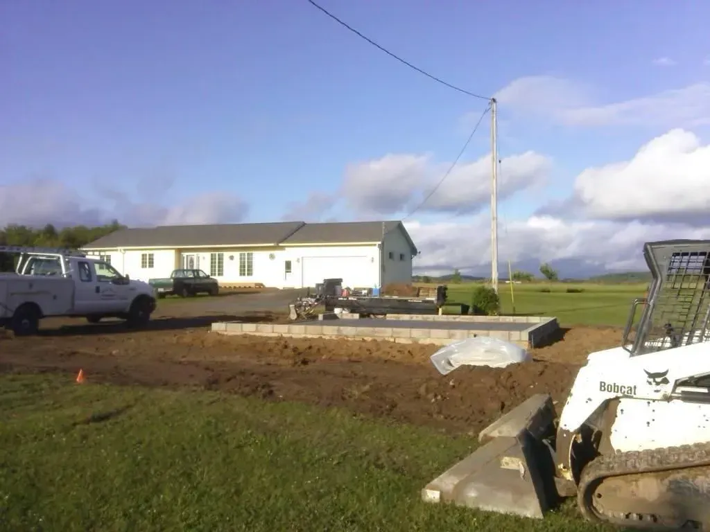 Construction site with a white work trailer, truck, and bulldozer in a rural field under a blue sky