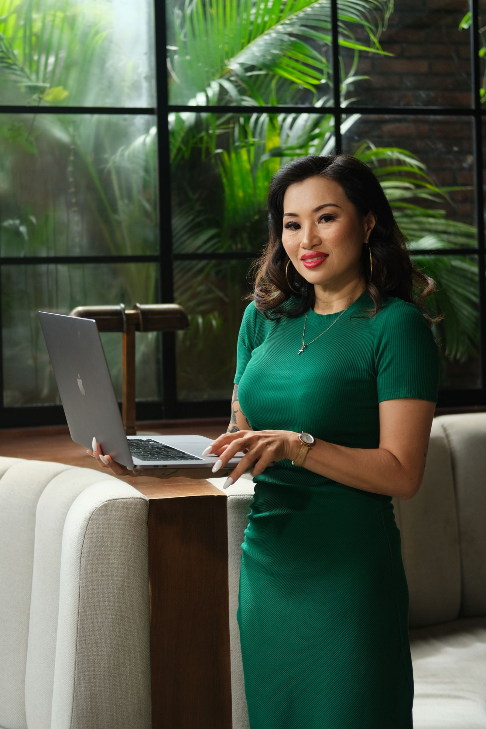 Woman in green dress works on laptop near a window with foliage.