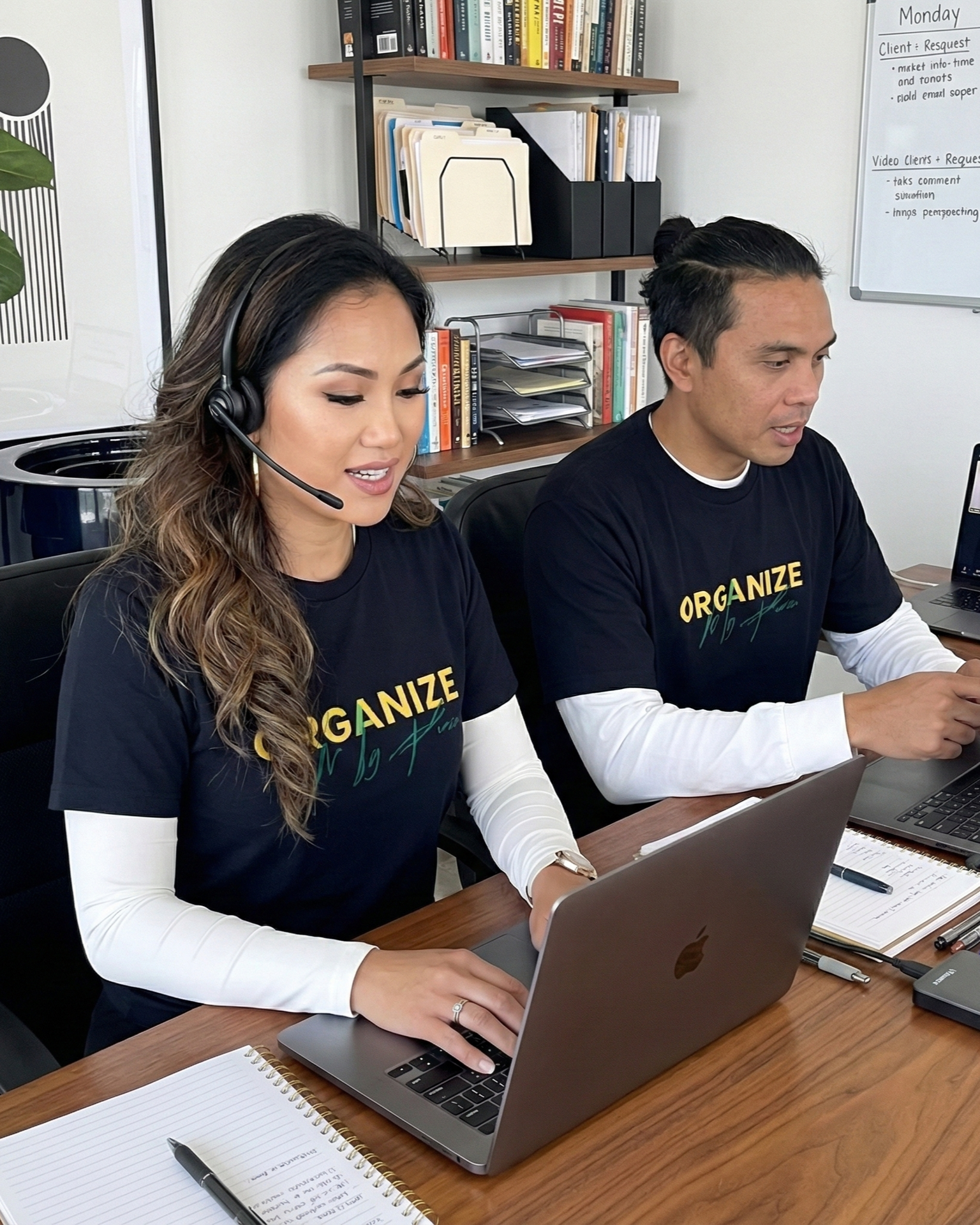 Two people at desks, working on laptops. One wears a headset. Both are wearing black shirts with