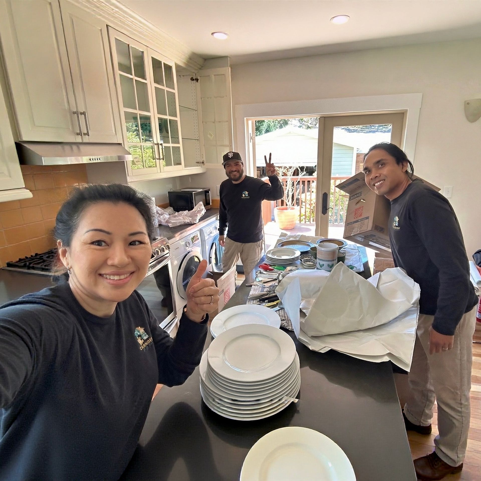 Movers in a kitchen; smiling, packing dishes. One gives a thumbs up, another a peace sign.