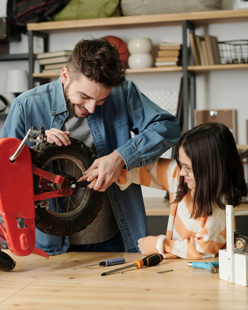 Man and child working on a red bicycle wheel; indoors, smiling and focused.