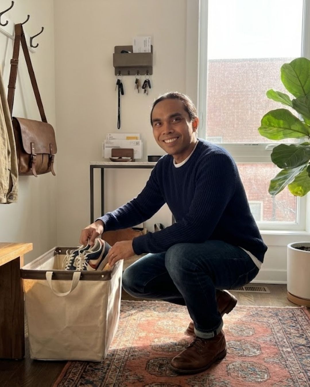 Man crouching, putting shoes in a canvas bin. Beside a window and a potted plant in a well-lit hallway.