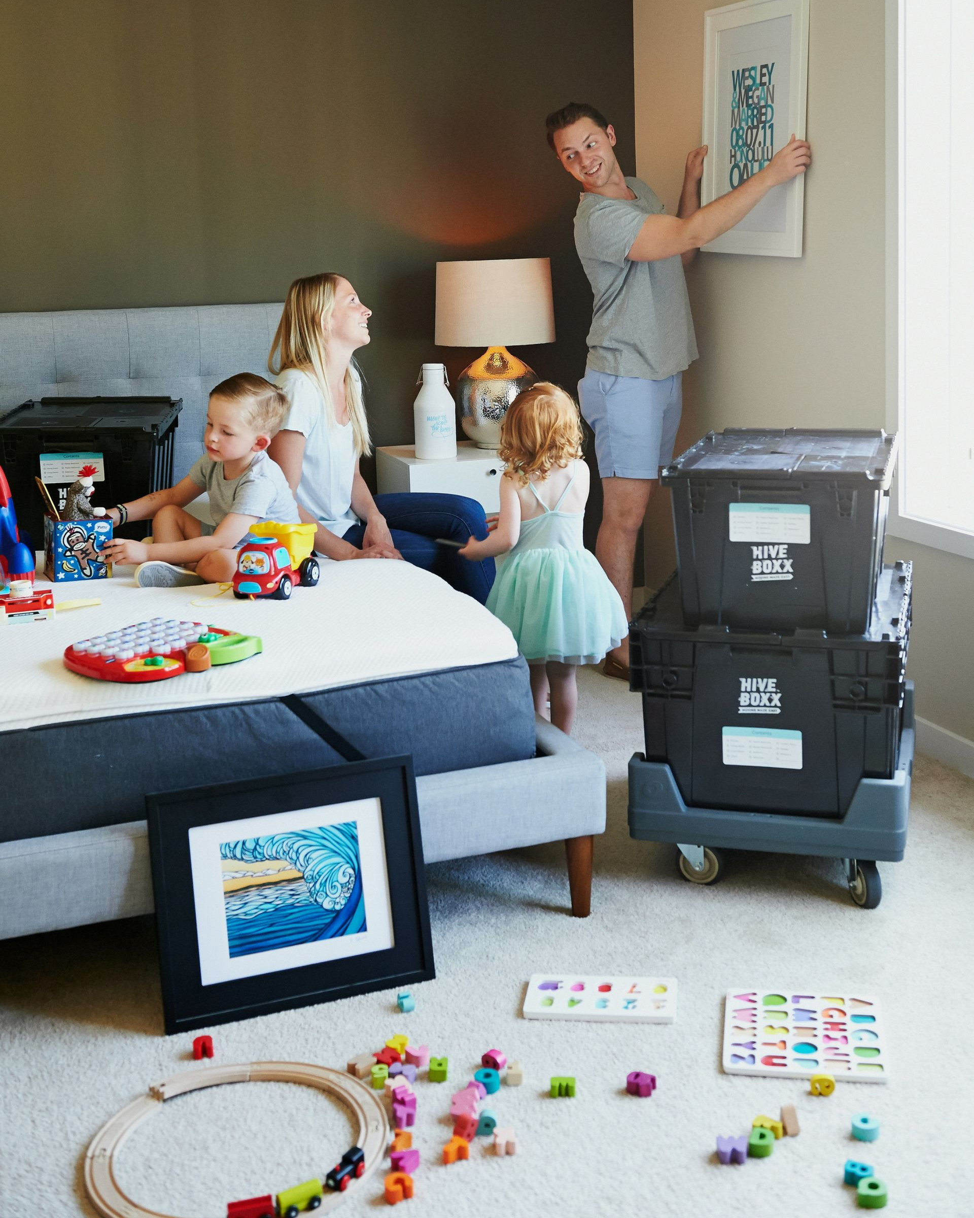 Family in a bedroom; man hangs art, woman and children sit on bed near moving boxes and toys.