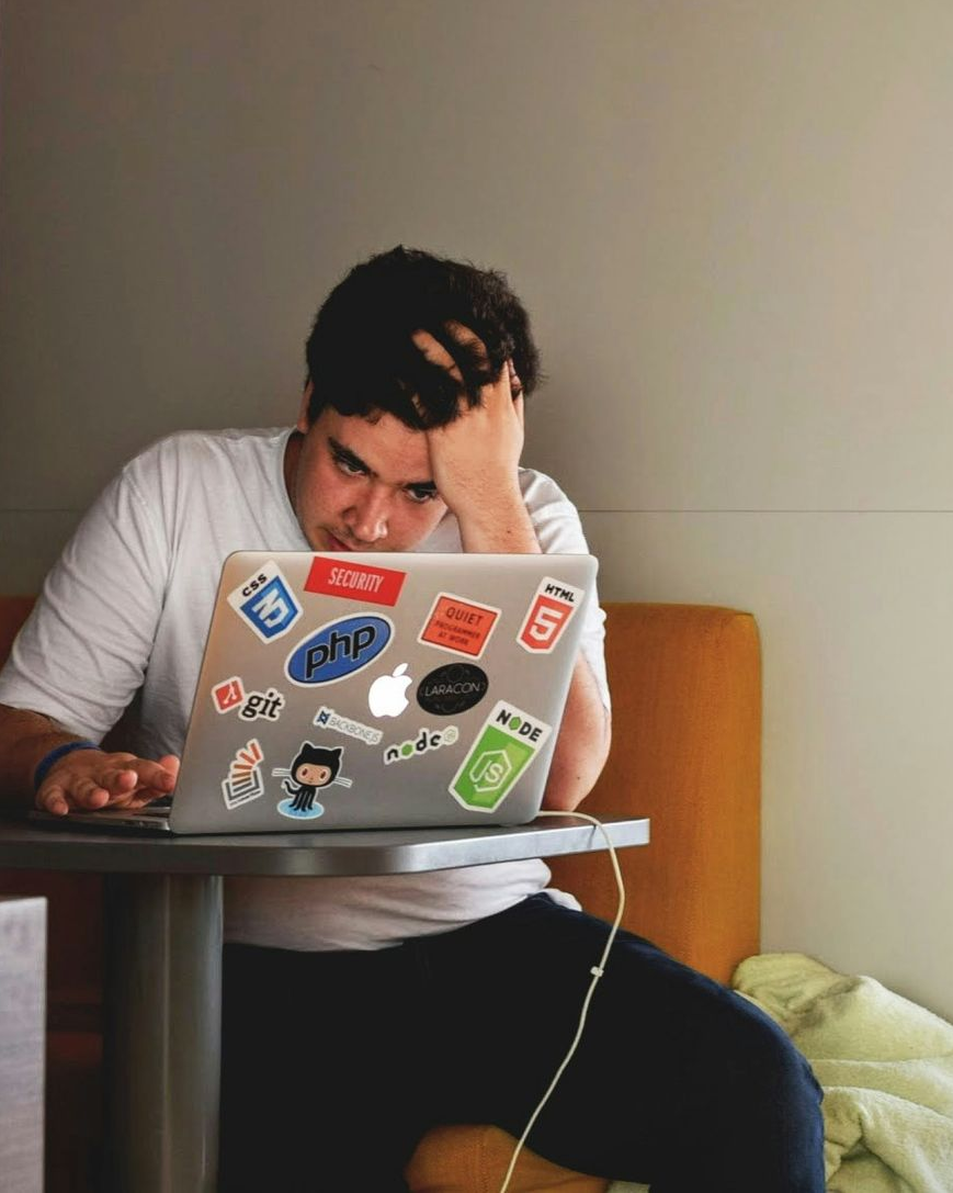 Man seated with laptop, looking stressed, hand on head. Silver laptop with stickers on small table, in orange booth.