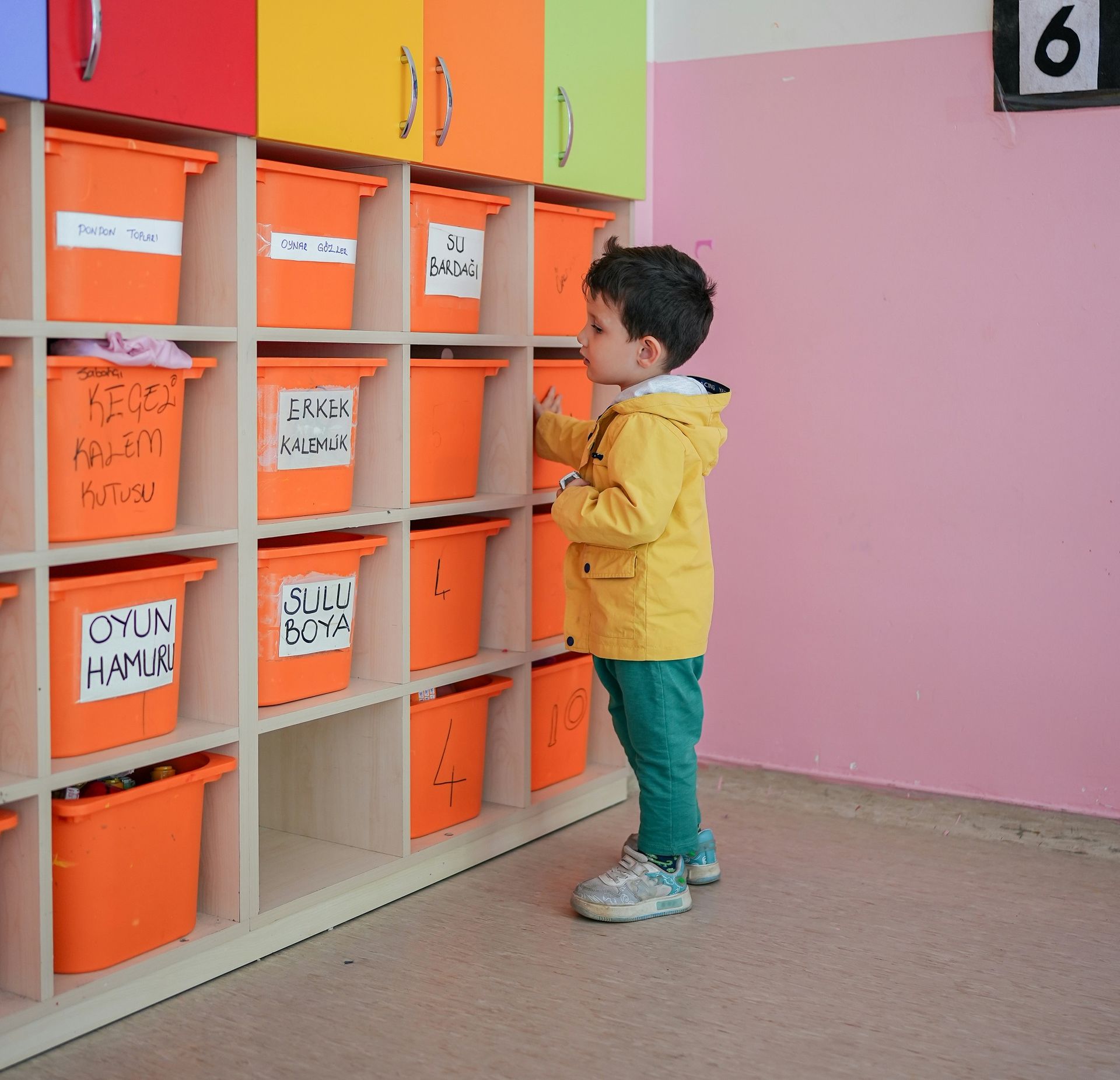 A young boy in a yellow jacket is standing in front of a shelf with orange bins that say oyun hamur