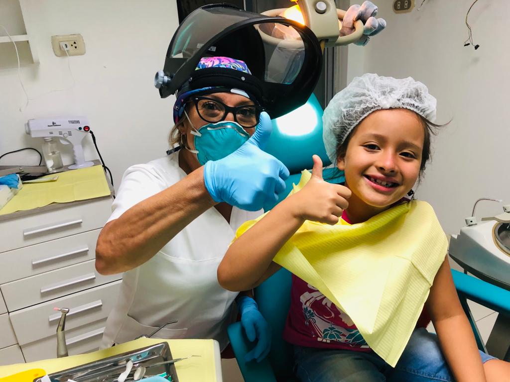 Una mujer le hace un gesto de aprobación con el pulgar hacia arriba a una niña sentada en una silla dental.