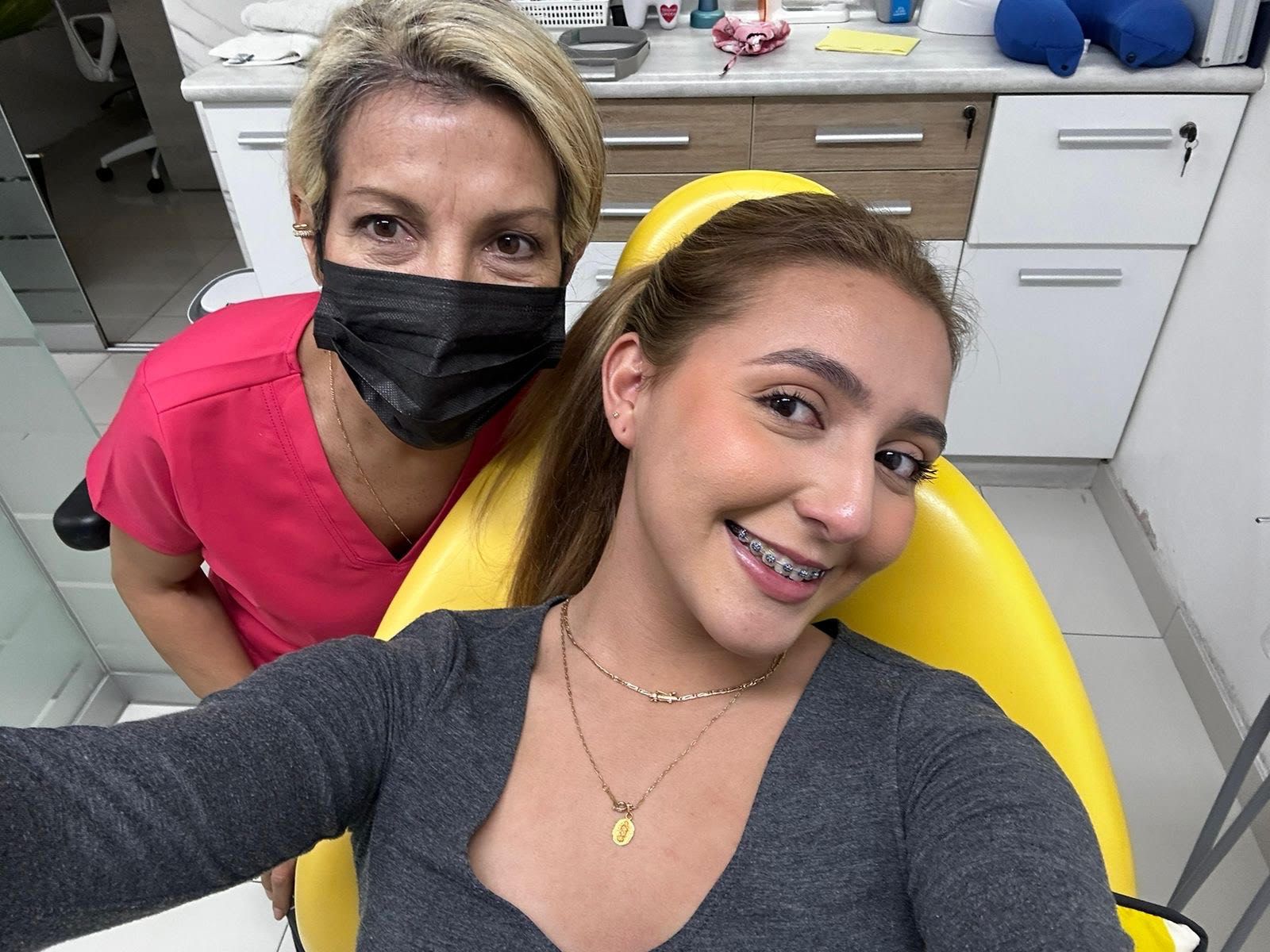 Una mujer está sentada en una silla dental al lado de una mujer que lleva una máscara.