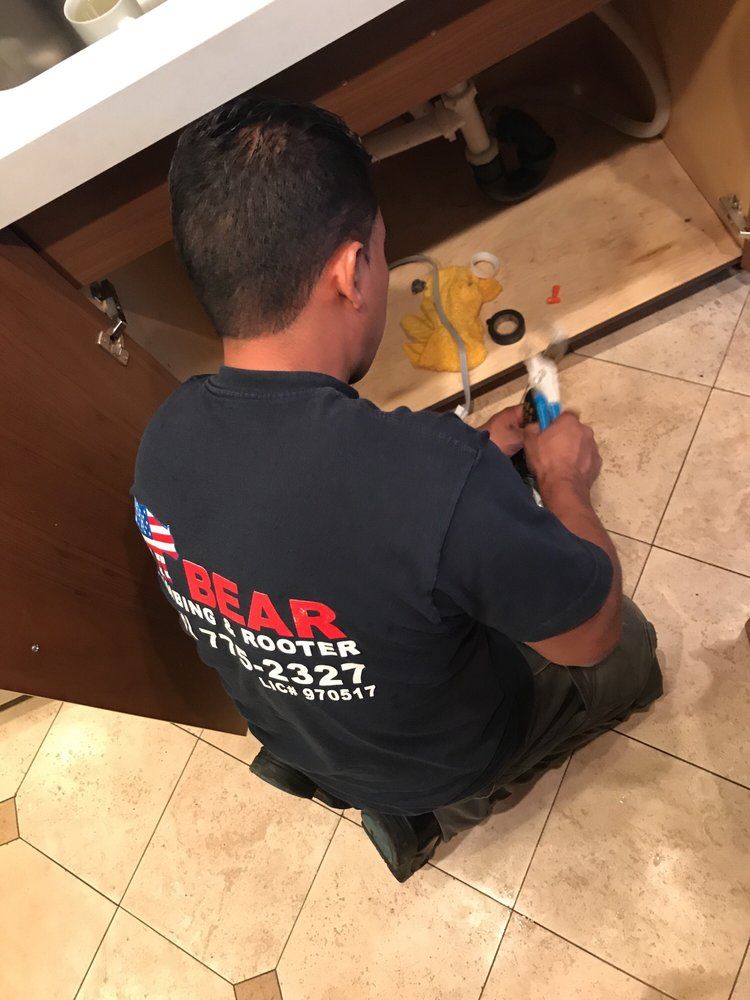 A plumber in a navy Bear Plumbing & Rooter shirt kneels while working under a kitchen sink.