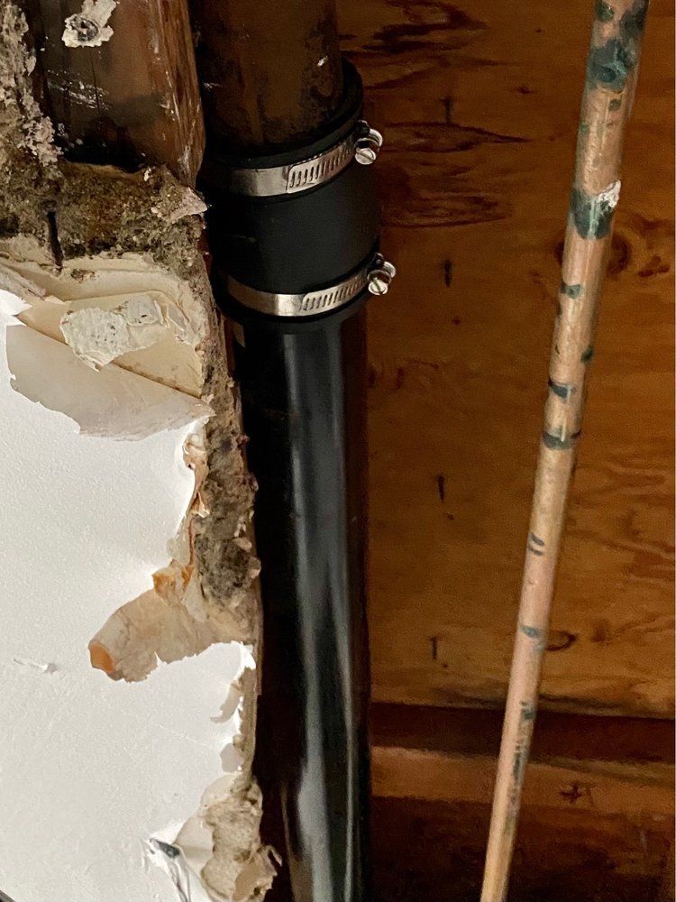A black rubber plumbing coupling with metal clamps connects a vertical metal pipe to a plastic pipe inside a wall.