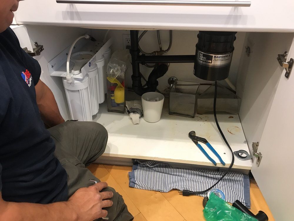 A plumber works under a kitchen sink, where a water filtration system and garbage disposal are installed.