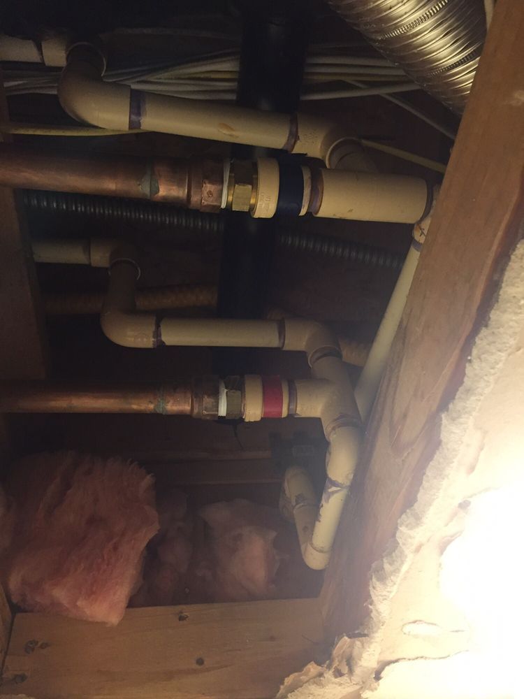 A black drain pipe connected by a metal rubber coupling to an elbow fitting in a wooden floor joist structure.