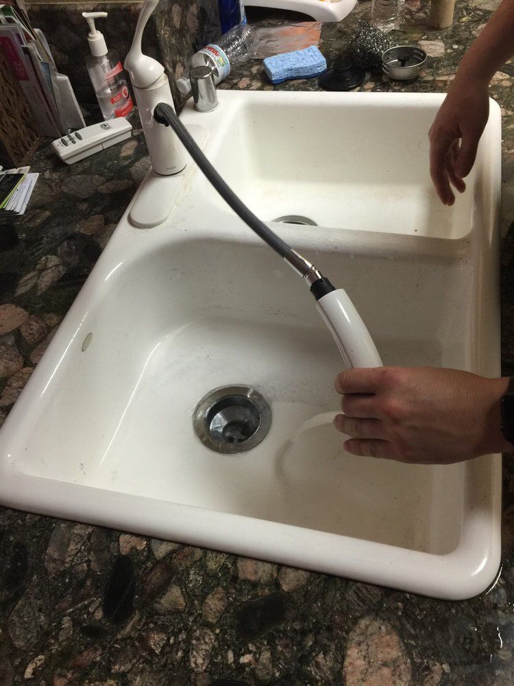 A person pulls the white, flexible sprayer hose out from a kitchen faucet over a double basin white sink.
